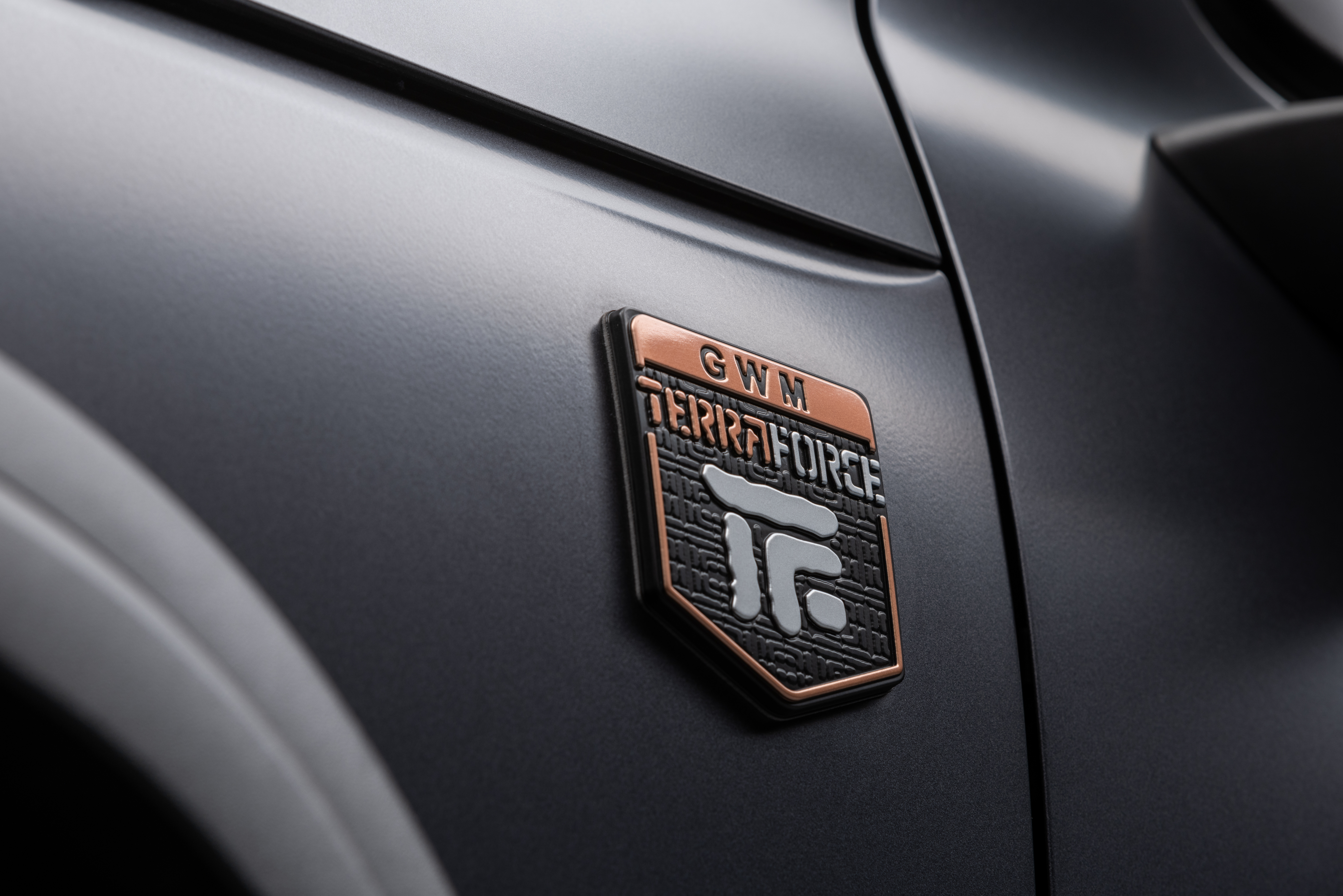 Emblema GWM Terraforce em tons de cobre e cinza, com fundo texturizado preto, fixado na lateral de um veículo cinza fosco