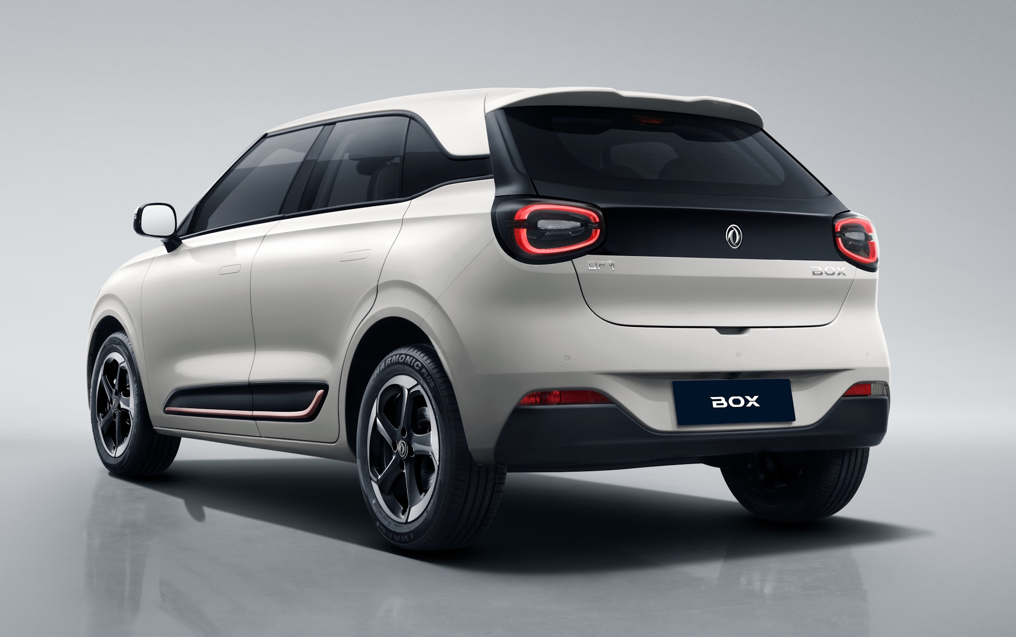 Carro elétrico branco, modelo SUV compacto, visto de traseira e lateral direita, com detalhes em preto na tampa do porta-malas e nas rodas. As lanternas traseiras são em formato de C invertido e a placa exibe BOX em branco sobre fundo azul escuro. O veículo está em um estúdio com fundo cinza claro e reflexo no chão