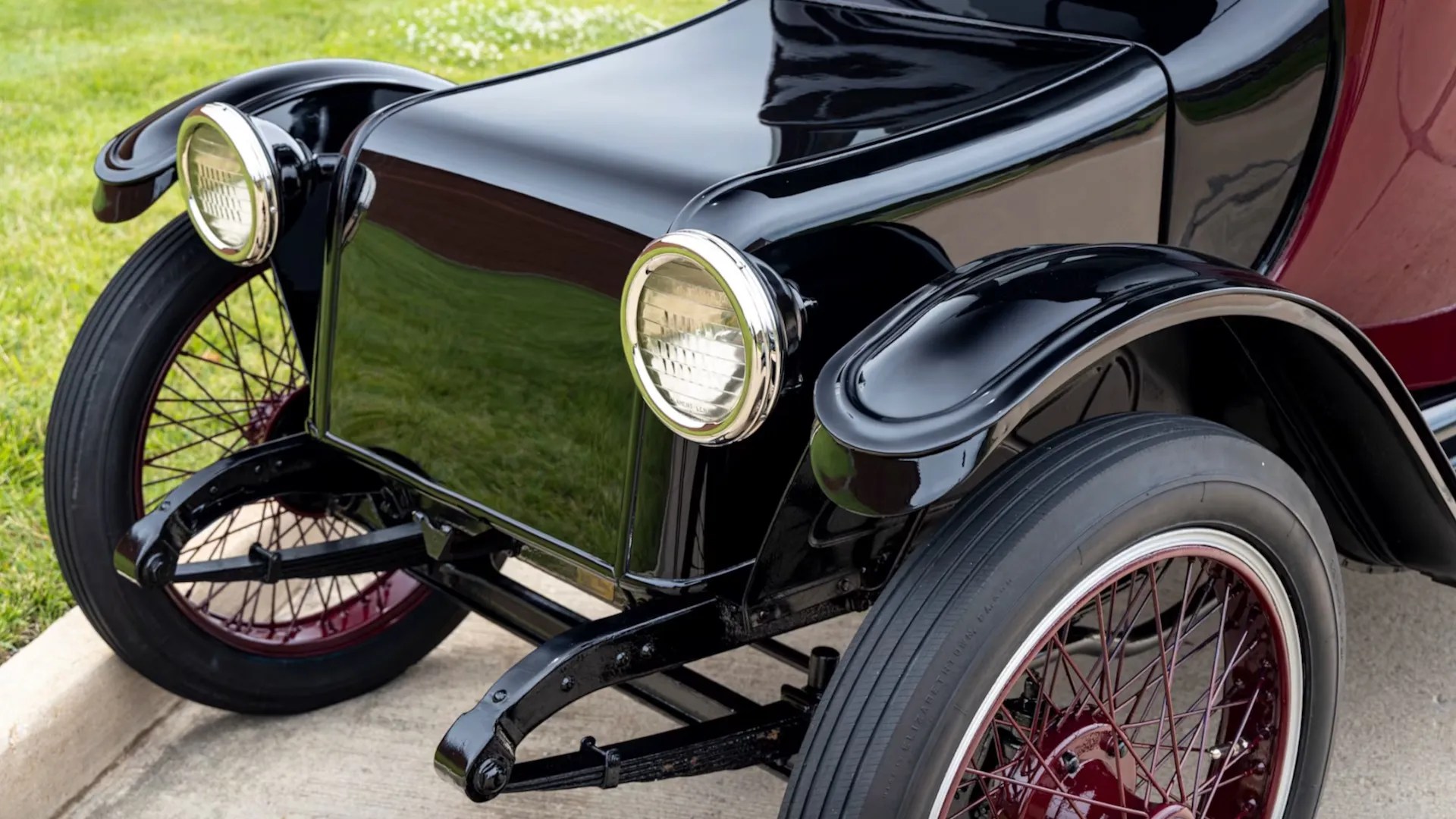 Parte frontal de um carro antigo preto e vinho, com para-lamas e faróis redondos cromados. As rodas têm aros e raios cor de vinho, e pneus pretos finos. O veículo está sobre um piso de concreto, com grama verde ao fundo
