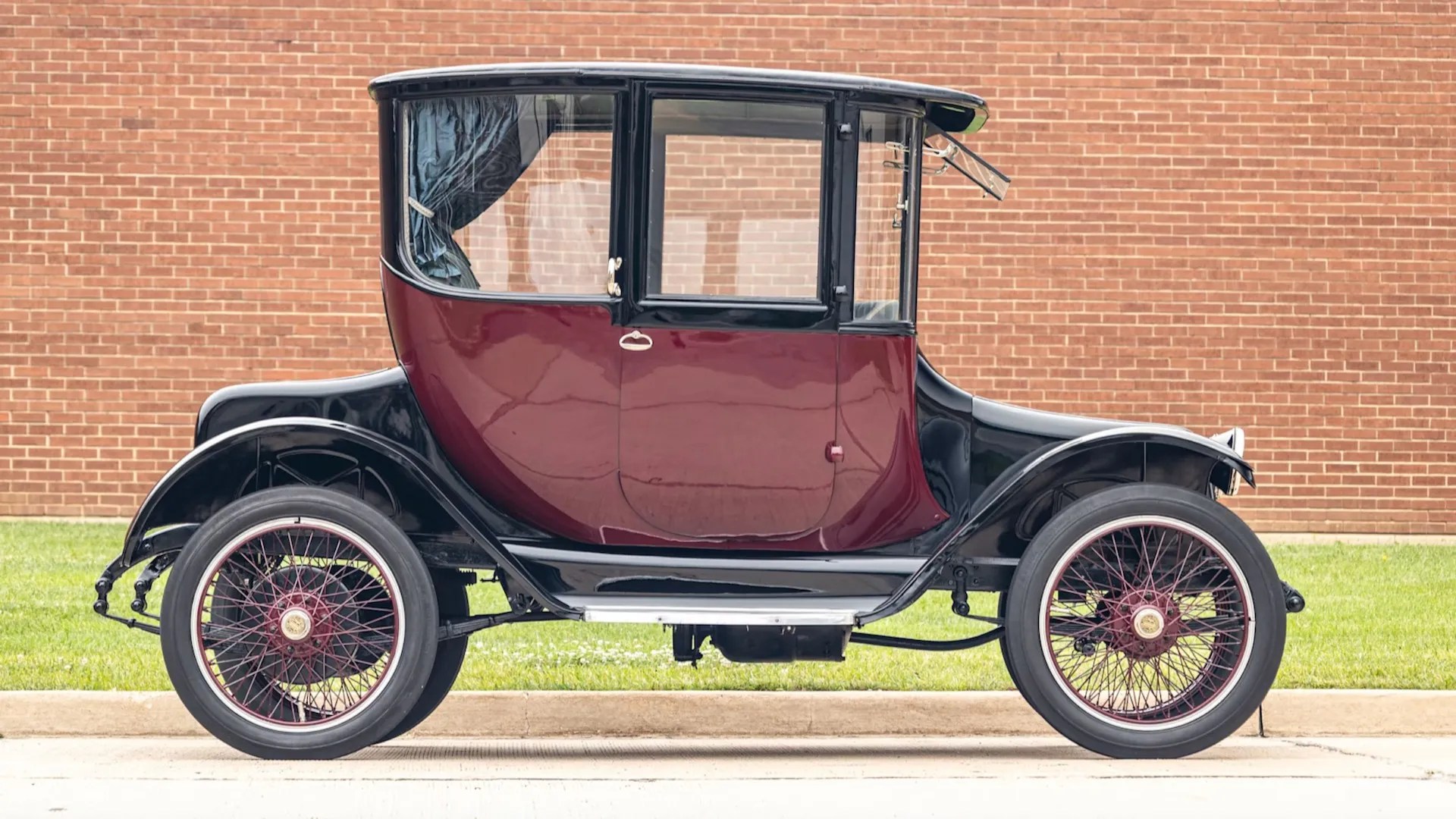 Carro antigo elétrico de 1914, modelo Detroit Electric, com carroceria preta e portas vinho, rodas raiadas com detalhes vermelhos, estacionado em frente a uma parede de tijolos e gramado