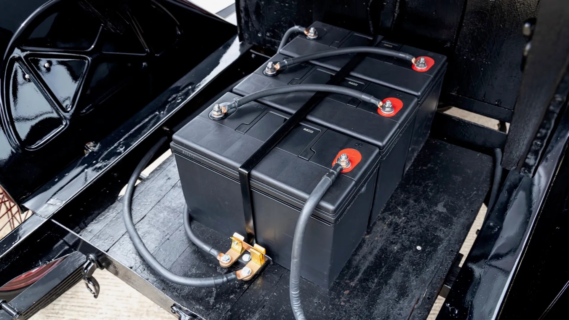 Três baterias automotivas pretas retangulares conectadas por cabos grossos, com terminais vermelhos e prateados, instaladas em um compartimento escuro de um veículo antigo