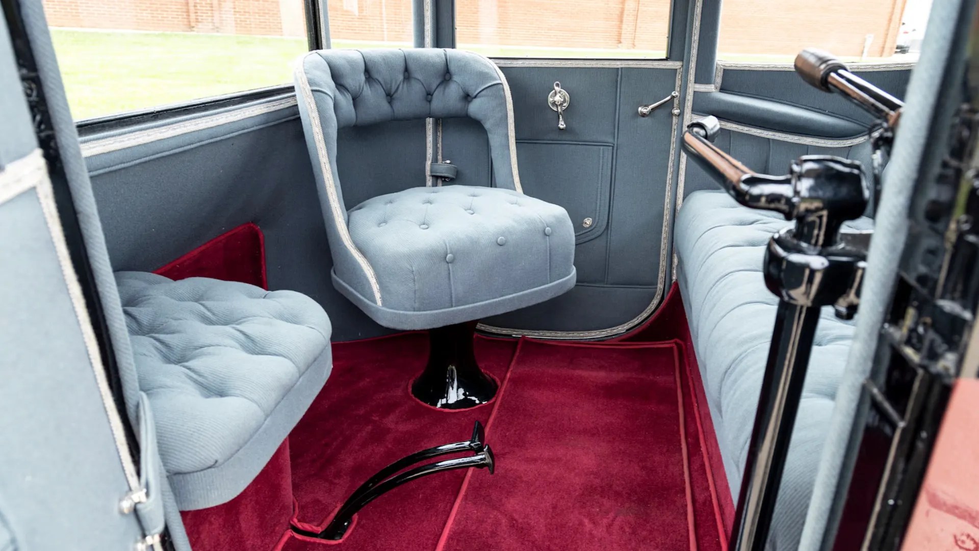 Interior de um carro antigo com estofamento cinza-azulado e carpete vermelho. Há três assentos visíveis: um giratório no centro, um menor à esquerda e um banco comprido à direita. As paredes são cinza-escuras com detalhes cromados e uma janela retangular. O volante e painel estão parcialmente visíveis à direita