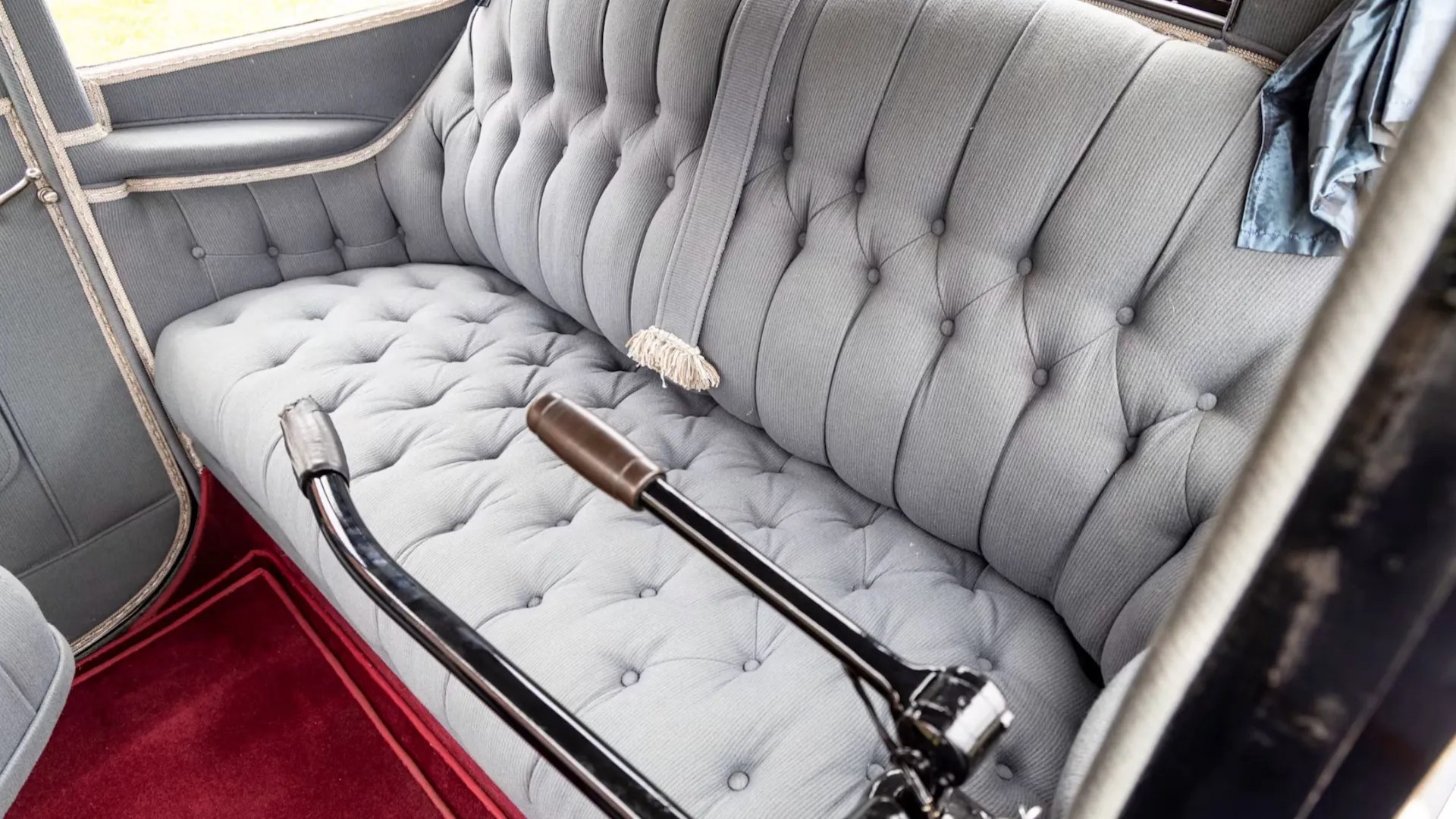 Interior de um carro antigo com banco traseiro estofado em tecido cinza claro, com botões e costuras formando losangos. O assoalho é coberto por um tapete vermelho e duas alavancas pretas com detalhes cromados e marrons se estendem do lado direito. Uma faixa de tecido com franjas brancas pende do encosto do banco.