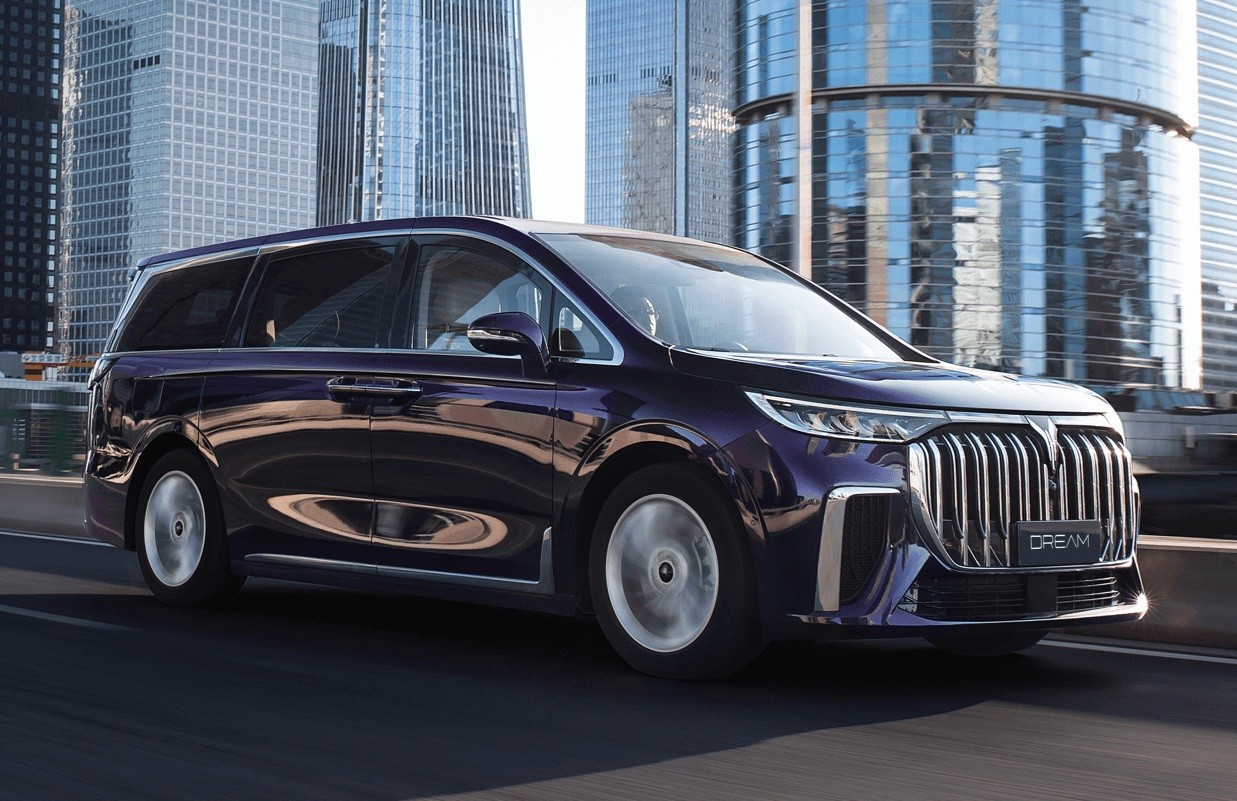 Minivan roxa escura, modelo Dream, em movimento numa via urbana, com prédios modernos de vidro ao fundo