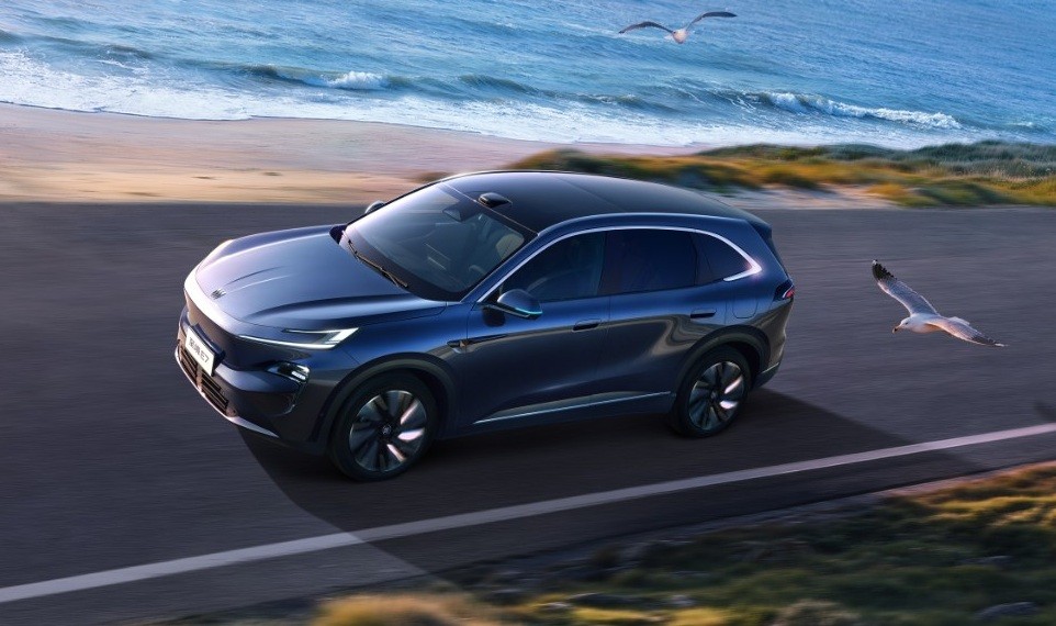 SUV azul-escuro em movimento numa estrada costeira, com o oceano e a areia ao fundo. Duas gaivotas voam, uma acima do carro e outra à direita, perto do chão