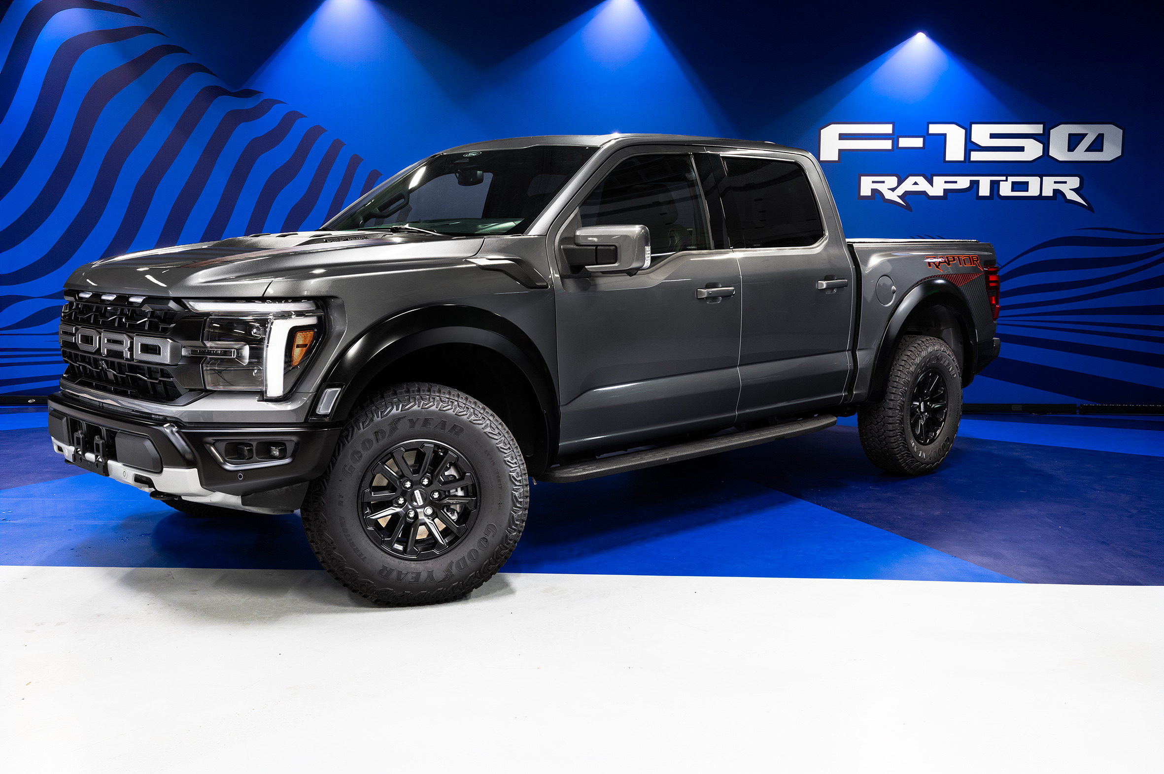 Picape Ford F-150 Raptor cinza escura, com pneus robustos e rodas pretas, exibida em um estúdio com iluminação azul e o logo F-150 RAPTOR na parede