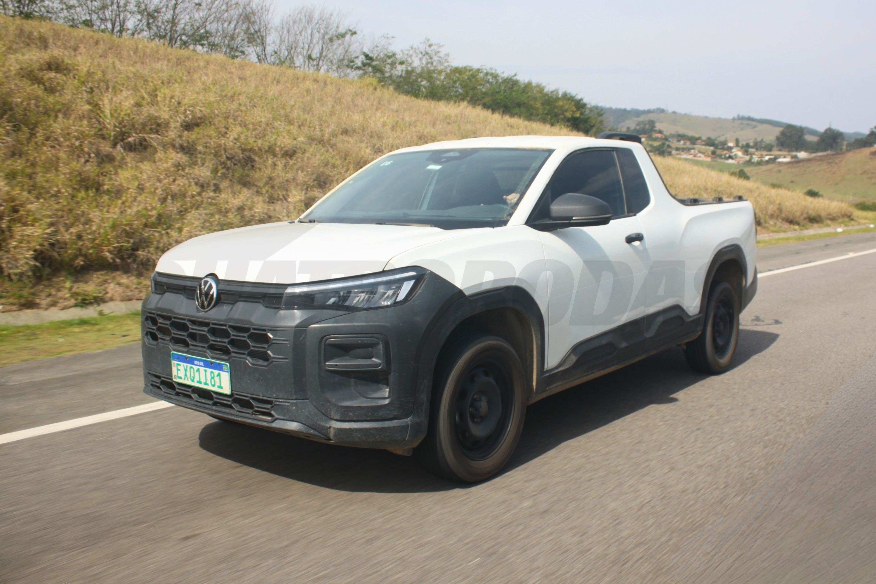 Picape branca Volkswagen com cabine simples e caçamba, rodando em estrada asfaltada. O veículo tem para-choque e molduras das rodas em plástico preto fosco, e placa de teste verde com a inscrição EXD1I81. Ao fundo, morros com vegetação seca e céu claro