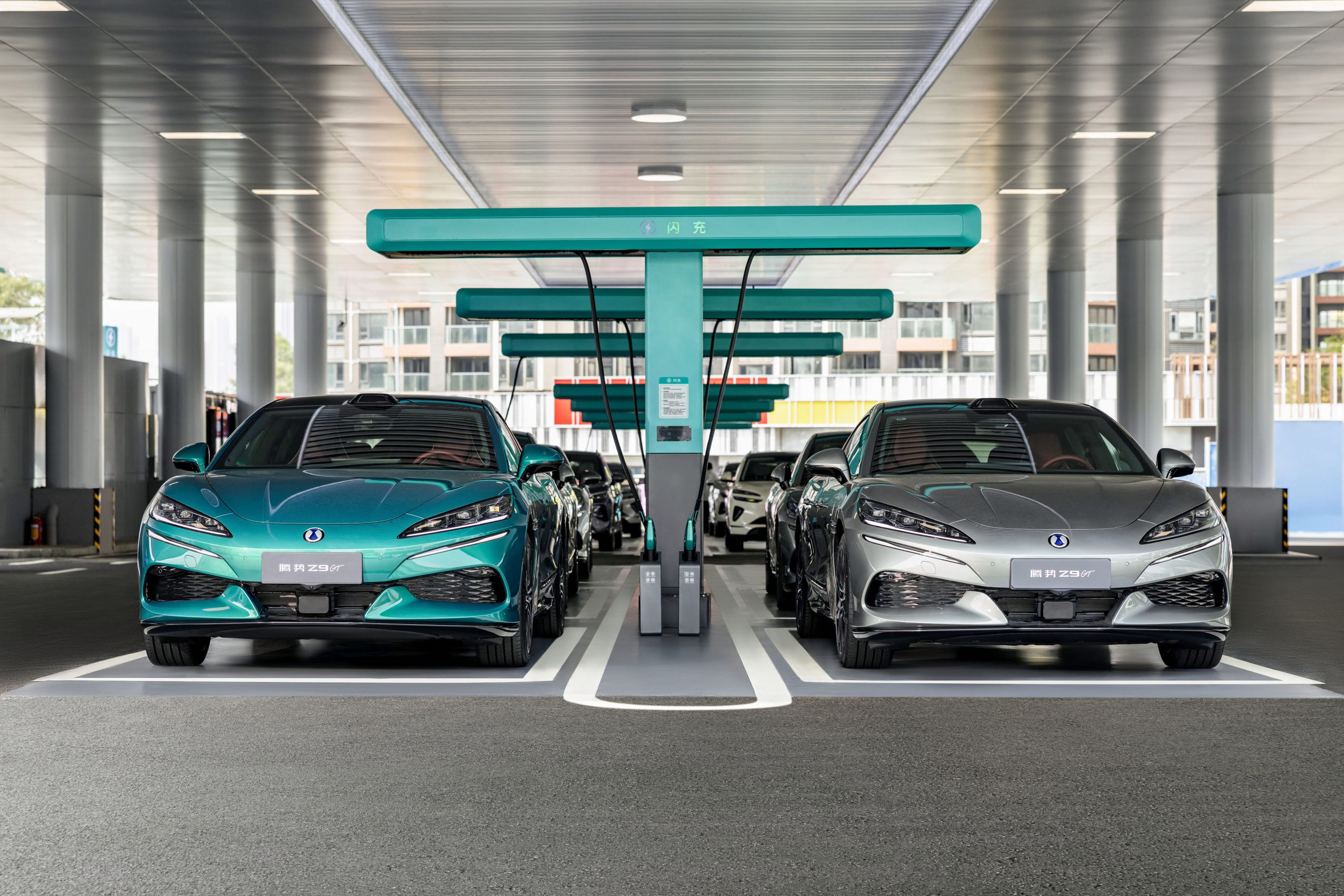 Dois carros elétricos, um azul-petróleo e um prata, estacionados lado a lado em uma estação de carregamento coberta, com vários outros veículos ao fundo