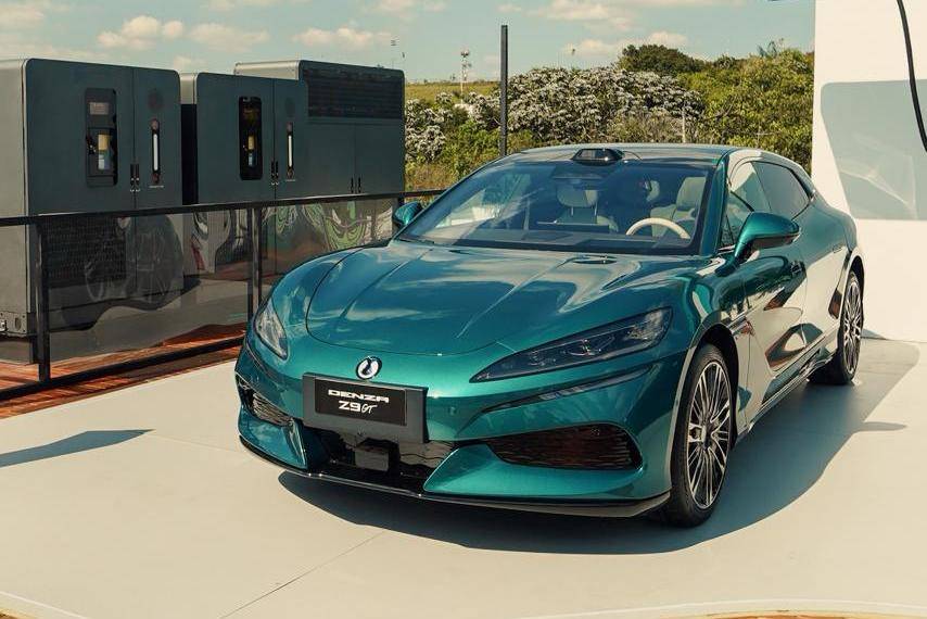 Flash Charging Brasil (1) Carro esportivo Denza Z9 GT verde-azulado metálico, estacionado em uma plataforma clara, com estações de carregamento cinzas ao fundo e vegetação