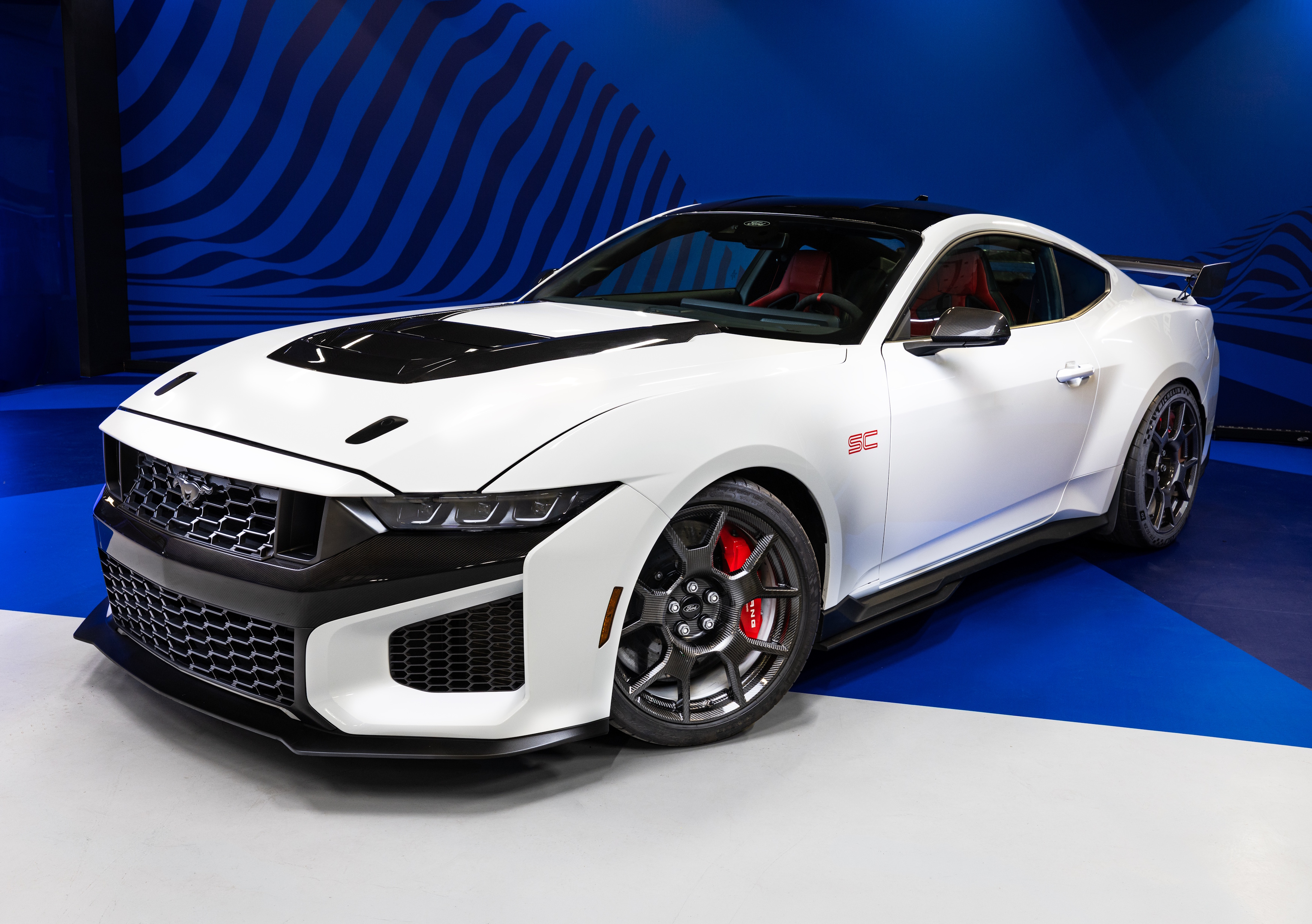 Carro esportivo branco com capô preto, rodas escuras e pinças de freio vermelhas, exibido em um piso cinza e azul com fundo azul ondulado