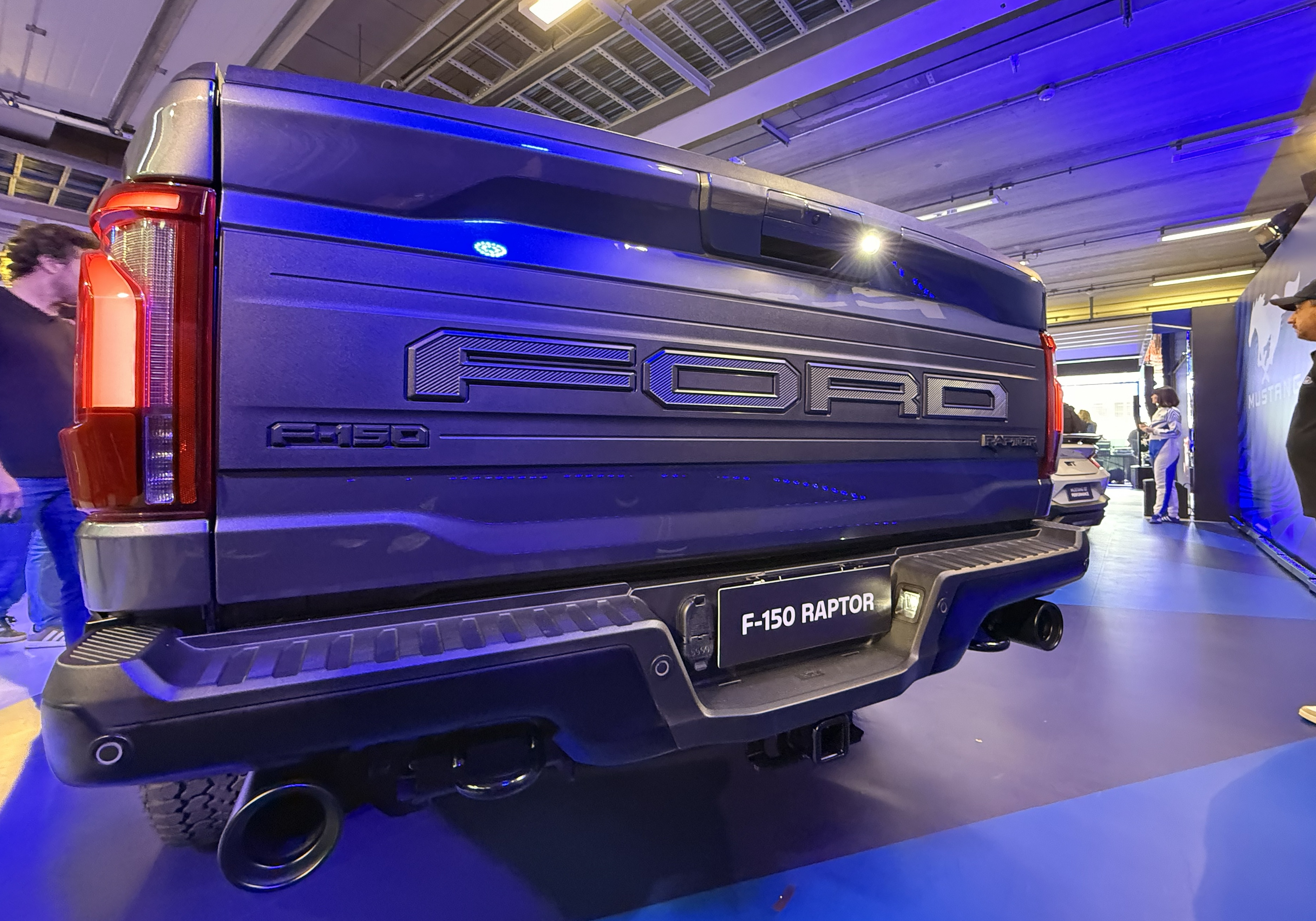 Traseira de uma picape Ford F-150 Raptor azul escura em um evento, com o nome FORD em relevo na tampa da caçamba e F-150 RAPTOR na parte inferior. Pessoas desfocadas aparecem ao fundo, e o ambiente é iluminado por luzes azuis