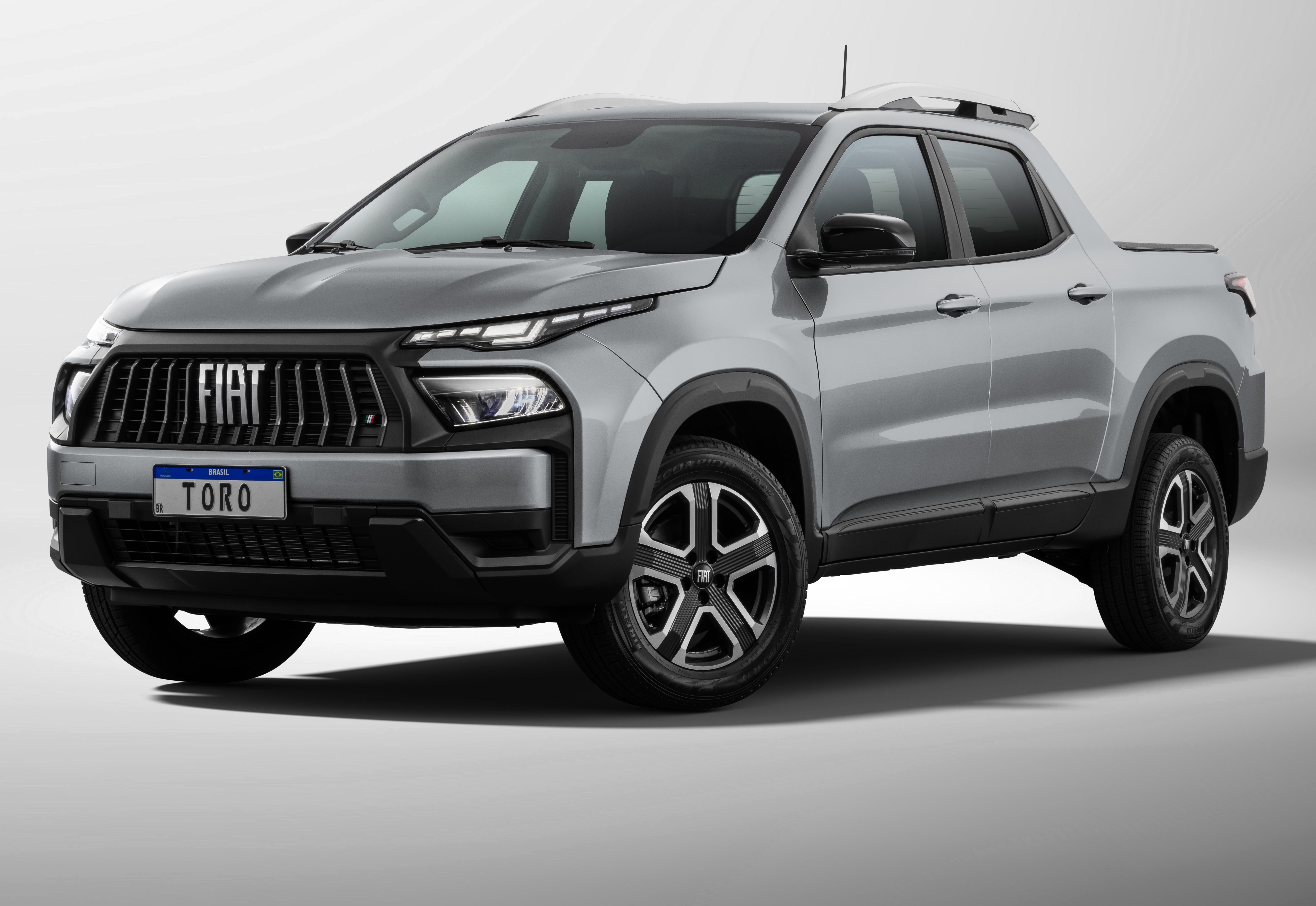 Picape Fiat Toro cinza-claro, com grade frontal preta e faróis de LED afilados, estacionada em um fundo branco. A placa frontal exibe TORO