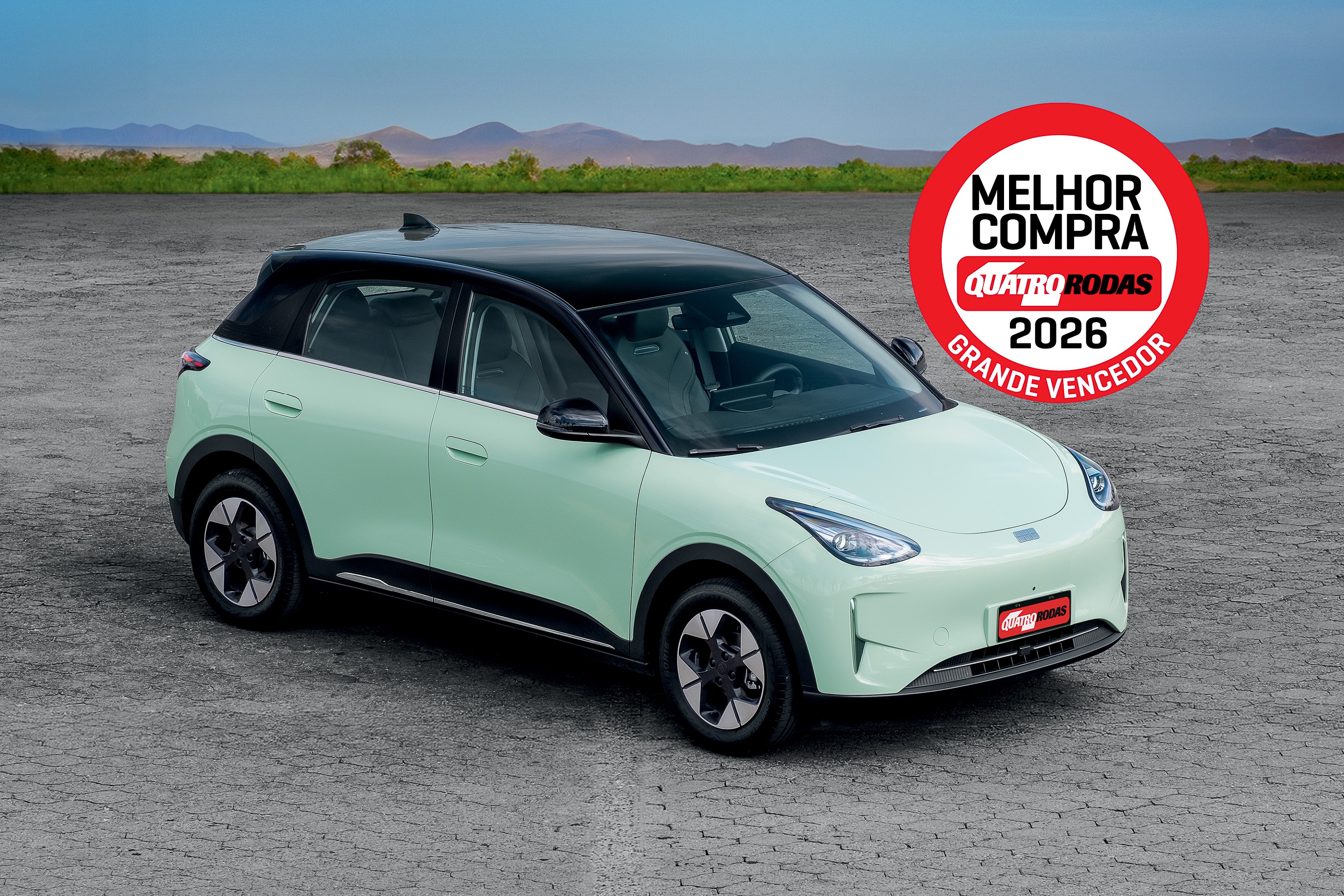 Carro el&eacute;trico verde-claro com teto preto, estacionado em piso cinza. No canto superior direito, selo vermelho Melhor Compra Quatro Rodas 2026 Grande Vencedor. Ao fundo, montanhas e vegeta&ccedil;&atilde;o