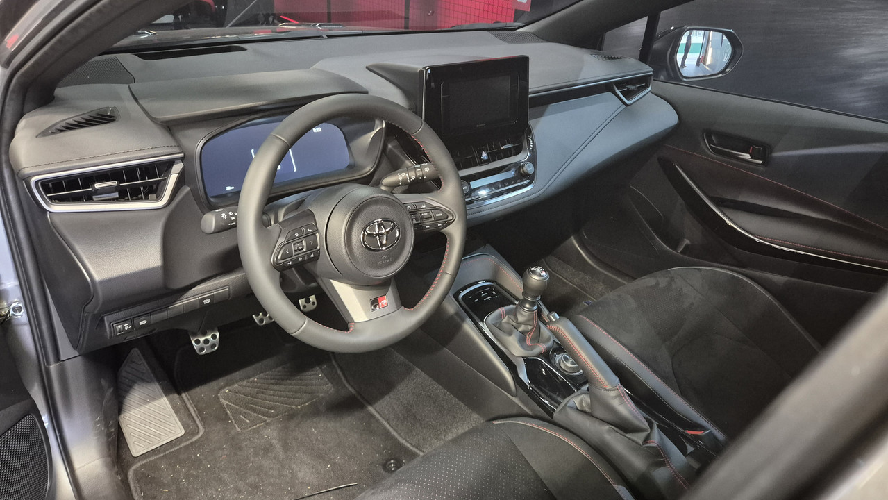 Interior de um carro esportivo cinza escuro, com volante multifuncional e logo Toyota, detalhes vermelhos no volante e câmbio manual. O painel possui tela digital e central multimídia, bancos de tecido escuro e pedais de metal. A porta do motorista está aberta, revelando o retrovisor lateral.