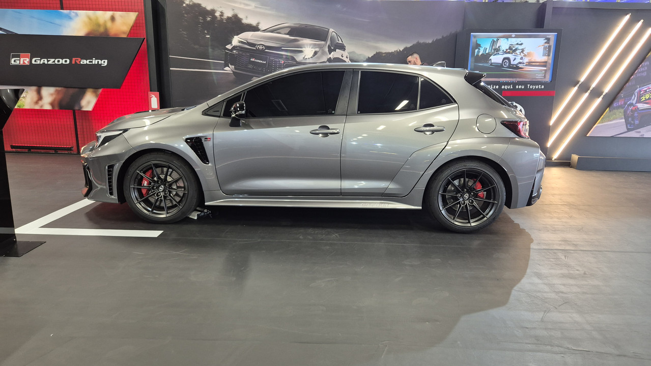Carro esportivo cinza-chumbo, modelo hatchback, com rodas pretas e pinças de freio vermelhas, estacionado em um showroom com painéis de fundo exibindo outros veículos e o logo GR Gazoo Racing
