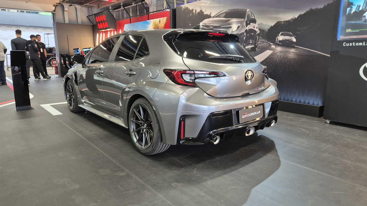 Carro cinza-chumbo, modelo hatchback, visto por tr&aacute;s e lateralmente, em um showroom. Possui rodas esportivas pretas, para-choque traseiro robusto com tr&ecirc;s sa&iacute;das de escape e um spoiler. Ao fundo, pessoas e um painel com imagens de carros em uma estrada