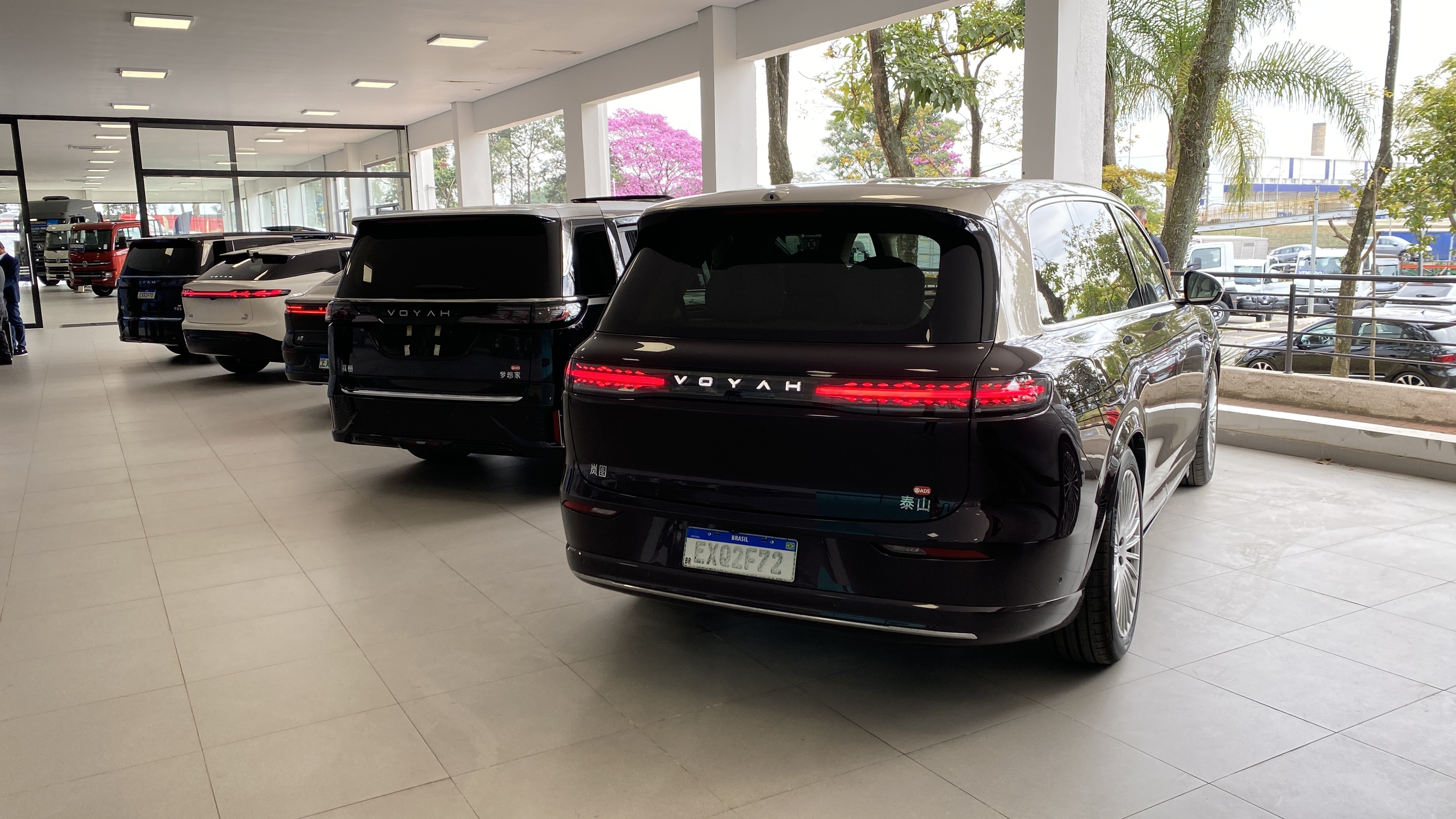 Vários carros de luxo, incluindo um SUV preto Voyah com placa brasileira, estacionados em uma concessionária com janelas grandes e árvores ao fundo