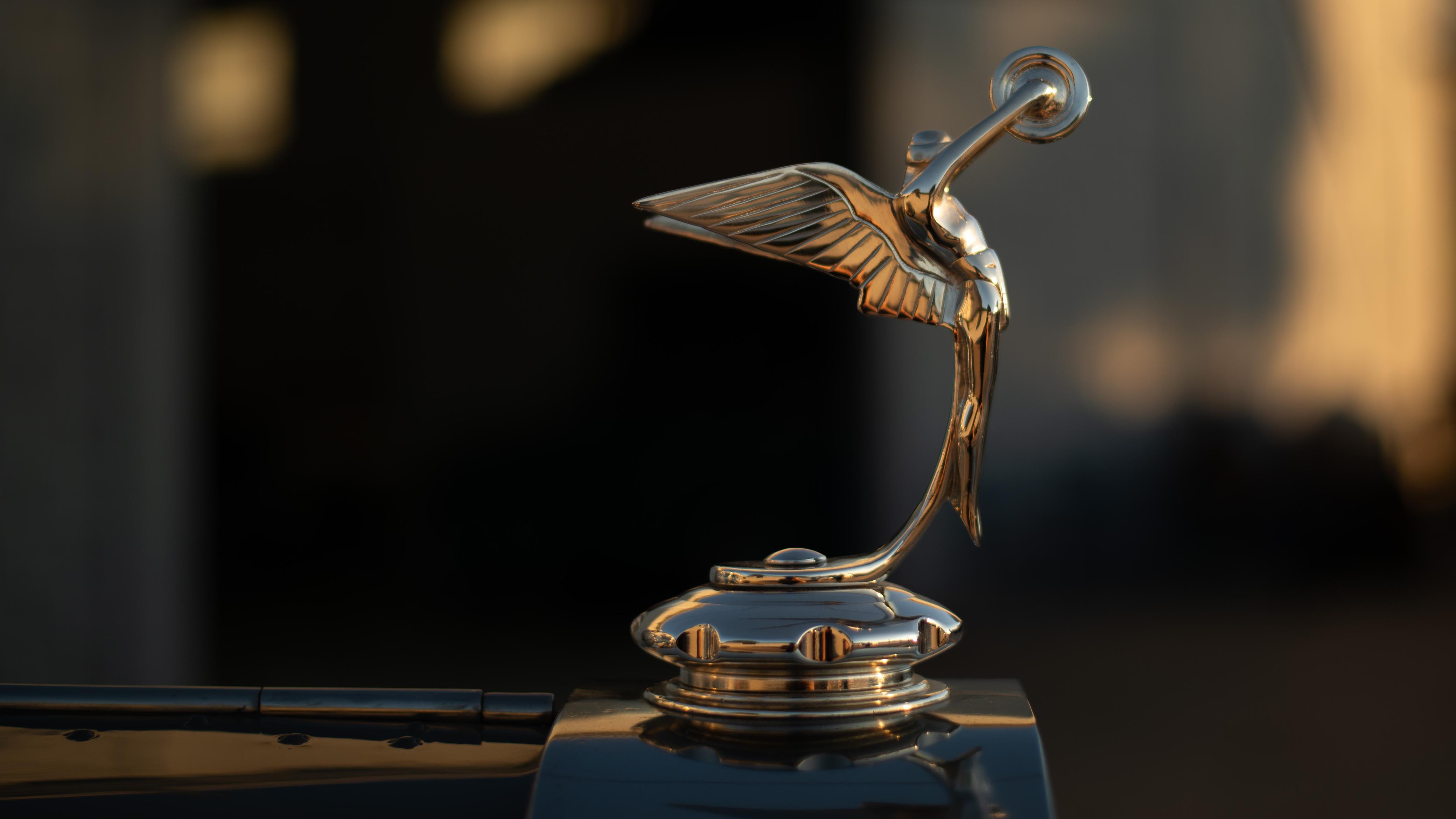 Estatueta dourada de uma figura feminina alada, com os braços estendidos para cima, adornando a frente de um carro antigo, com fundo escuro e reflexos dourados