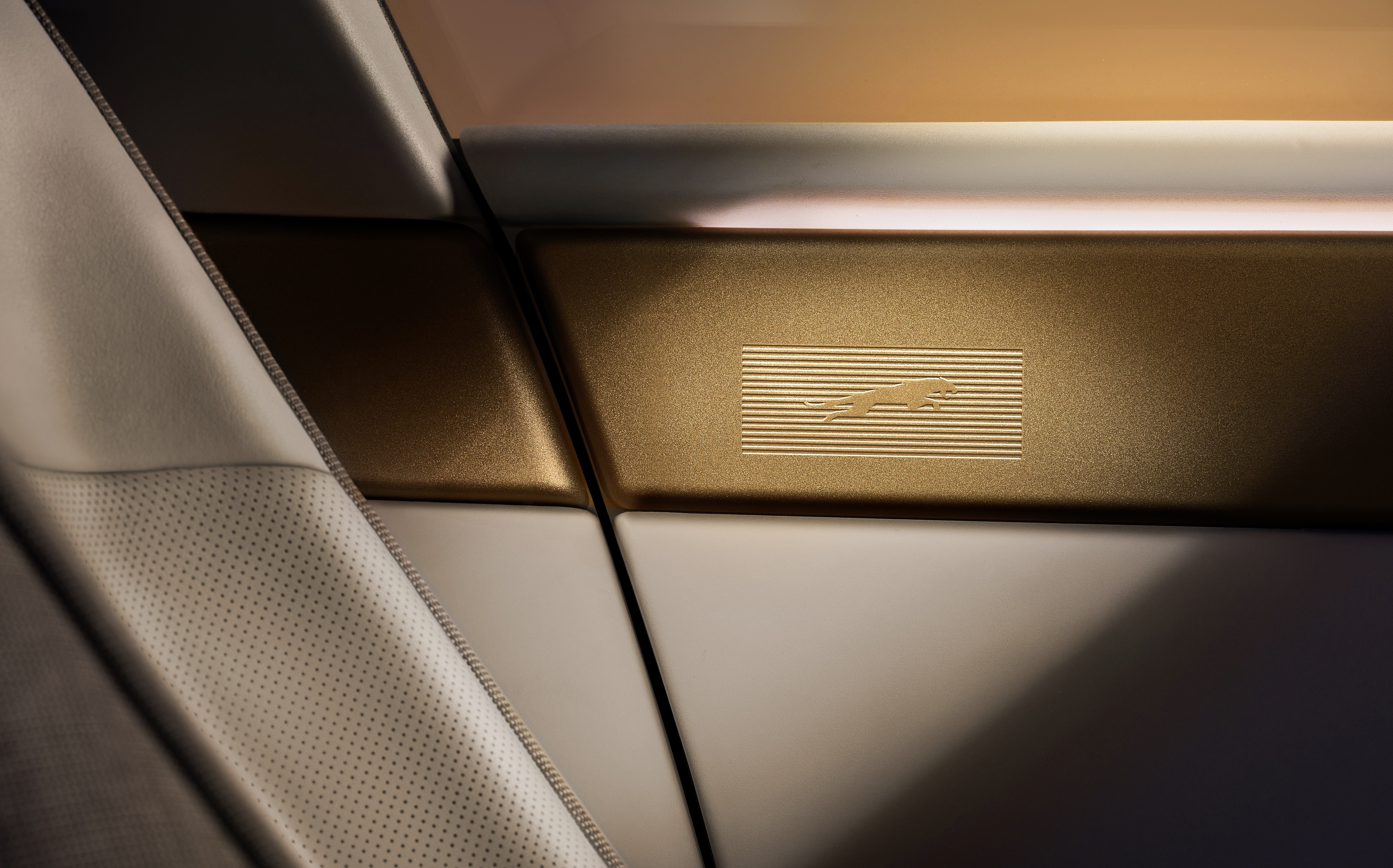 Detalhe do interior de um carro com acabamento em tons de branco e dourado. À esquerda, um assento de couro branco perfurado. No centro, uma faixa horizontal dourada com o logotipo de um jaguar em relevo sobre linhas horizontais finas. A iluminação cria um contraste de luz e sombra, destacando a textura dos materiais