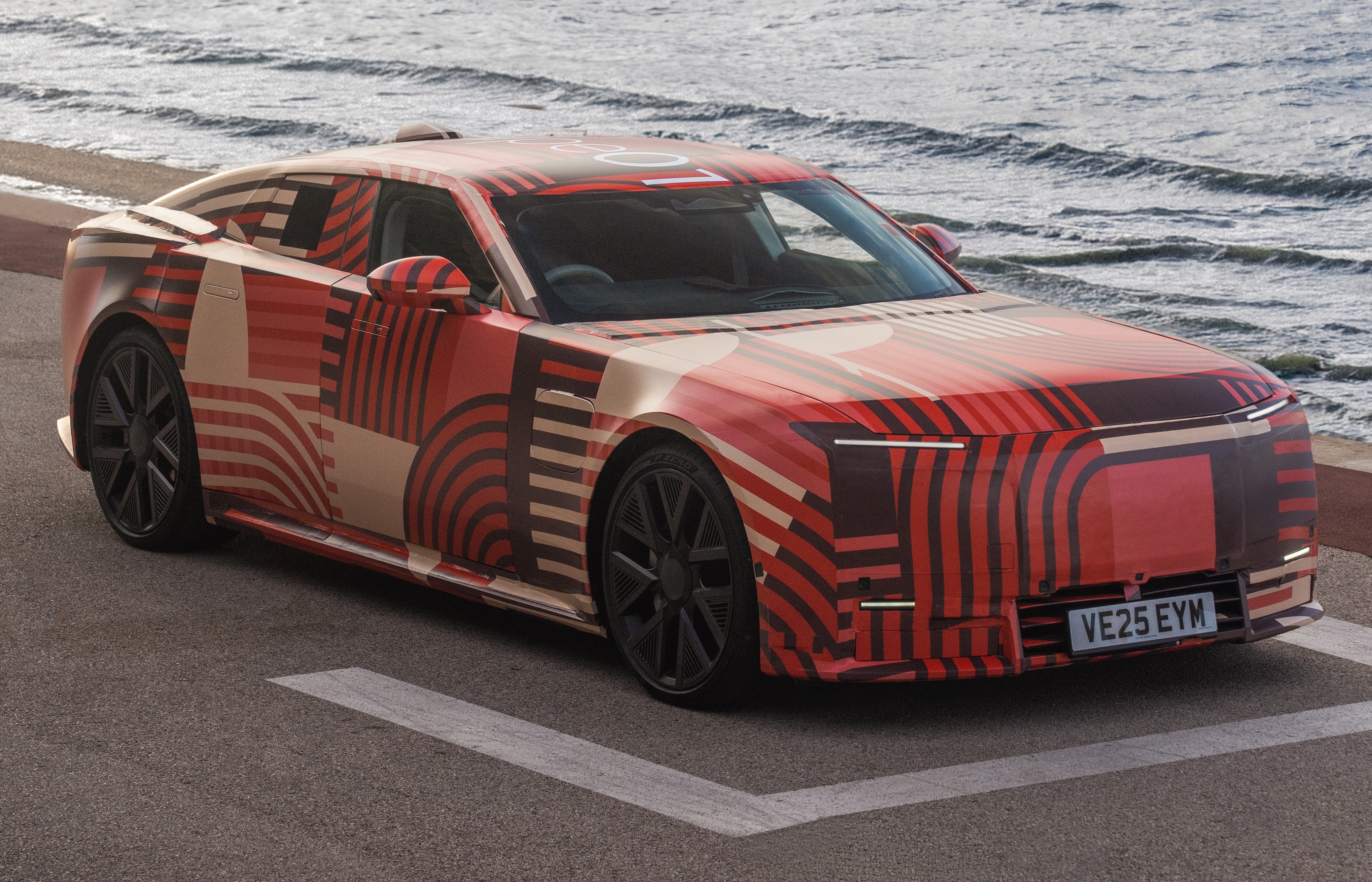 Carro protótipo camuflado com padrões geométricos em tons de vermelho, bege e preto, estacionado em uma rua asfaltada com o mar ao fundo