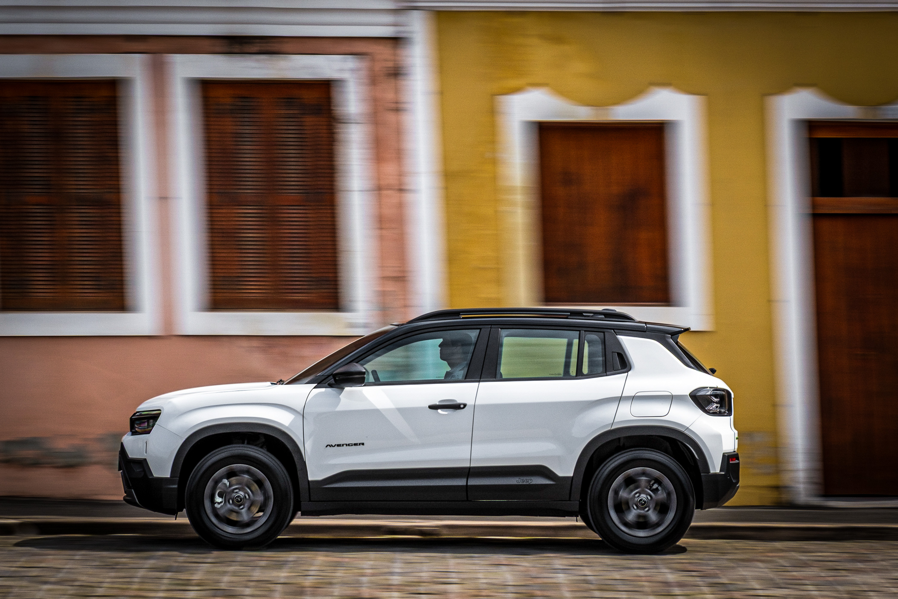 SUV branco com teto preto em movimento, visto de lado, passando por uma rua de paralelepípedos. Ao fundo, edifícios coloridos com janelas e portas de madeira escura, borrados pelo movimento