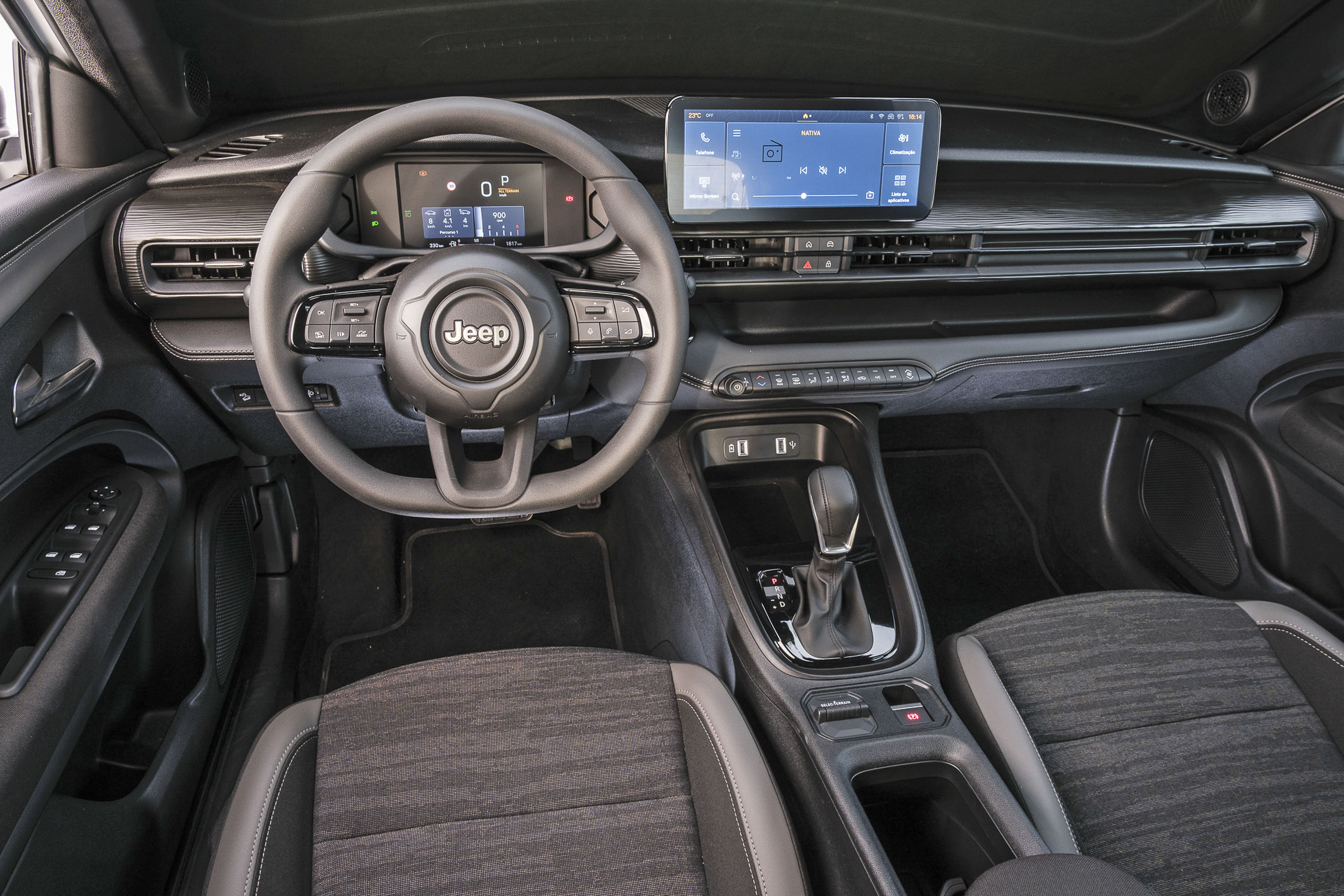 Interior de um carro Jeep, mostrando o volante com o logo da marca, painel digital, tela multimídia central com interface azul, console central com câmbio automático e bancos de tecido escuro com detalhes cinza