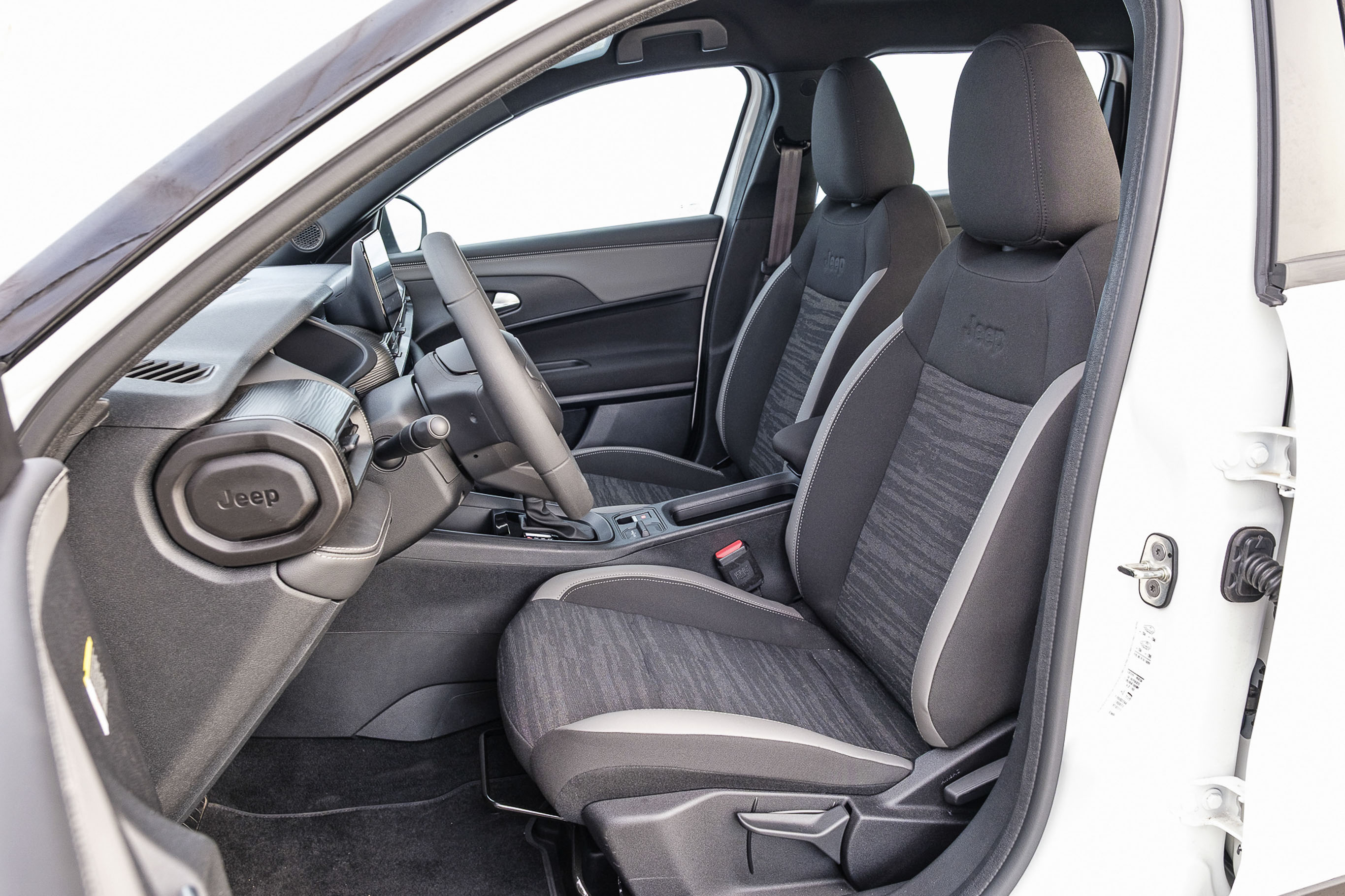 Interior de um carro branco, mostrando o banco do motorista e do passageiro da frente em tecido cinza escuro com detalhes em cinza claro e estampa abstrata. O volante e o painel são pretos, com o logotipo Jeep visível no painel lateral esquerdo. A porta do motorista está aberta, revelando o batente branco