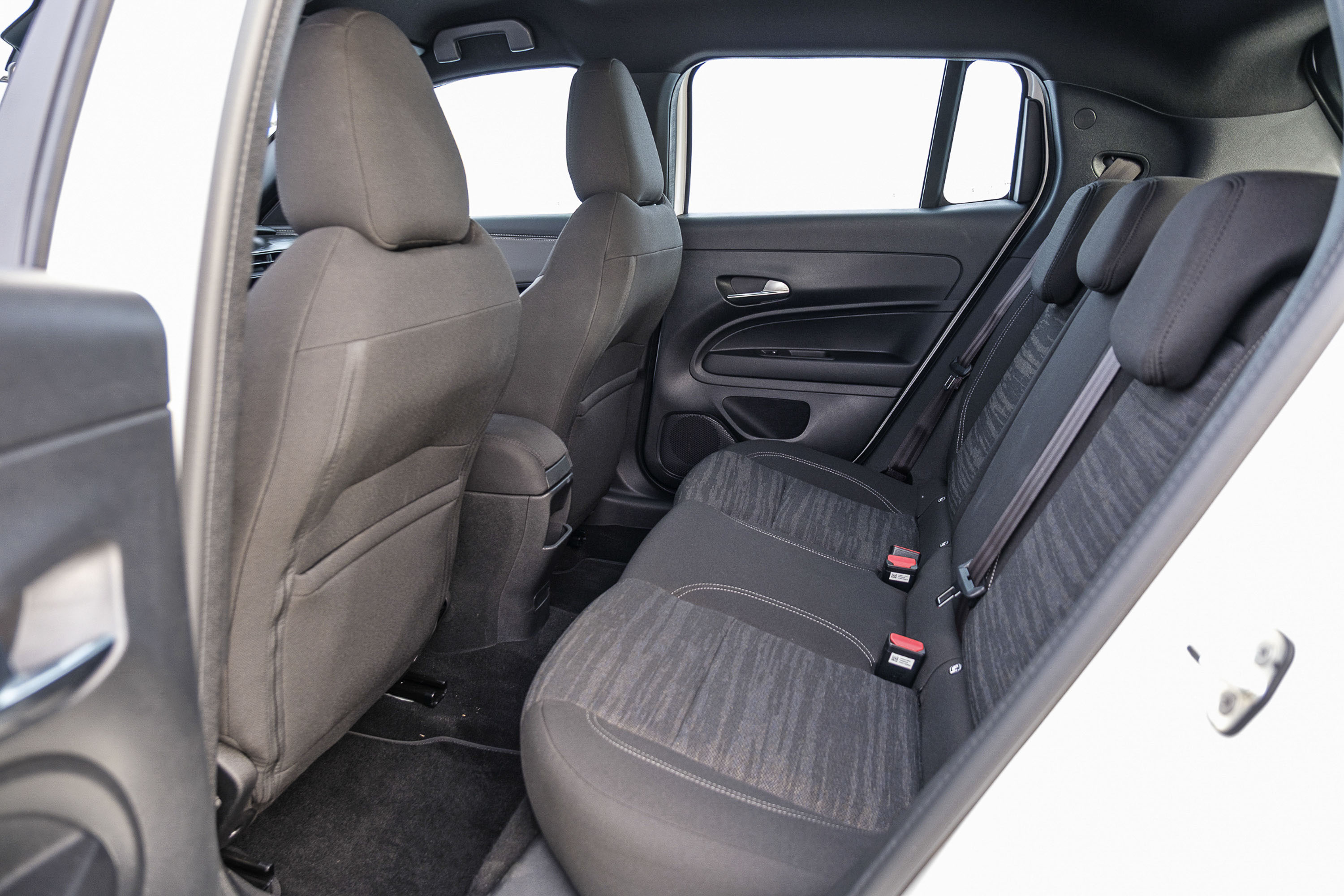 Interior de um carro branco, mostrando os bancos traseiros e parte dos dianteiros. Os bancos são cinza escuro, com estofamento de tecido texturizado nos assentos traseiros e encostos de cabeça pretos. Cintos de segurança com fivelas vermelhas estão visíveis. As portas e o teto são pretos, e as janelas mostram um fundo branco claro. A imagem é tirada de dentro do carro, com a porta traseira direita aberta.