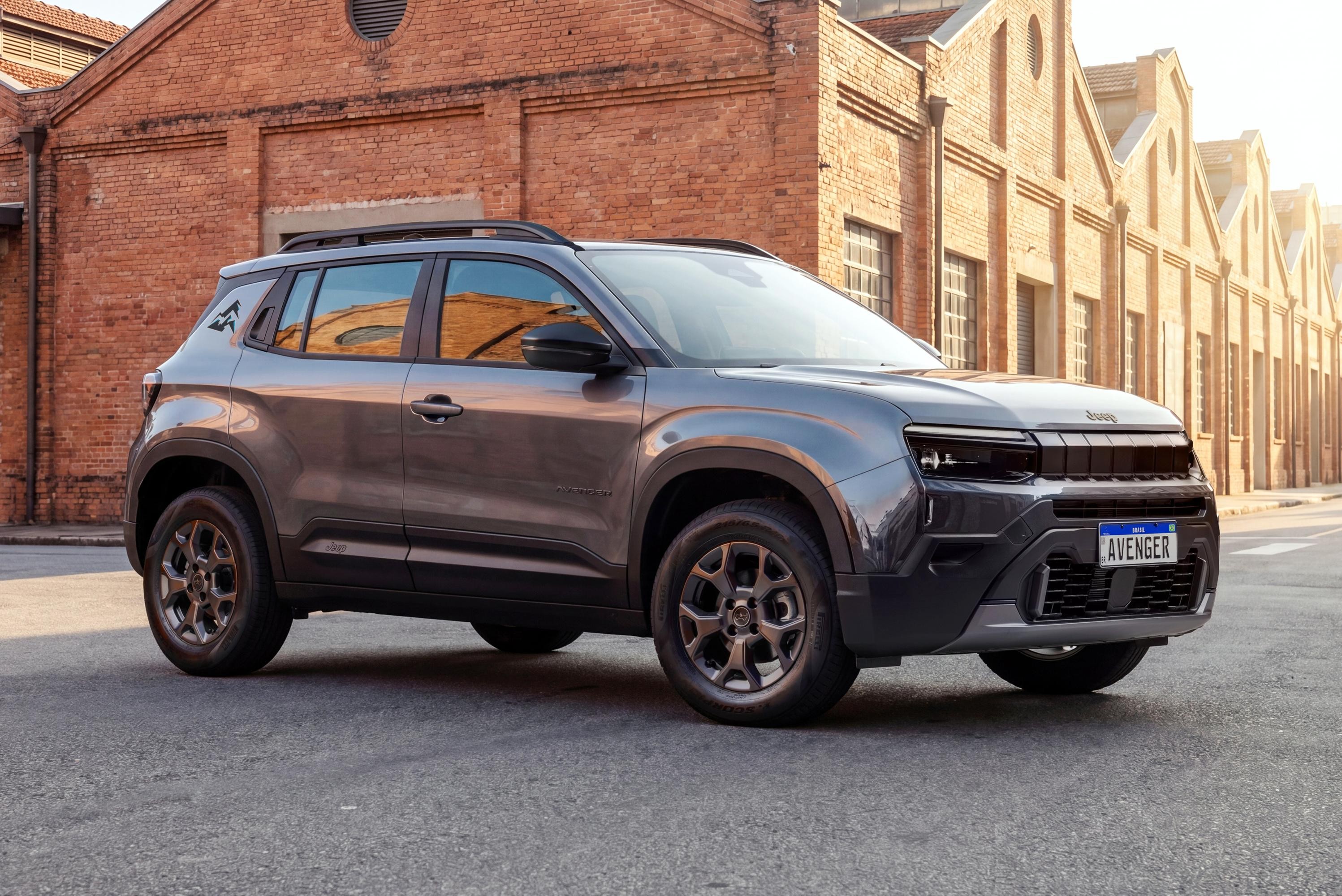 SUV Jeep Avenger cinza escuro estacionado em rua de paralelepípedos, com prédios de tijolos avermelhados ao fundo e luz solar forte à direita