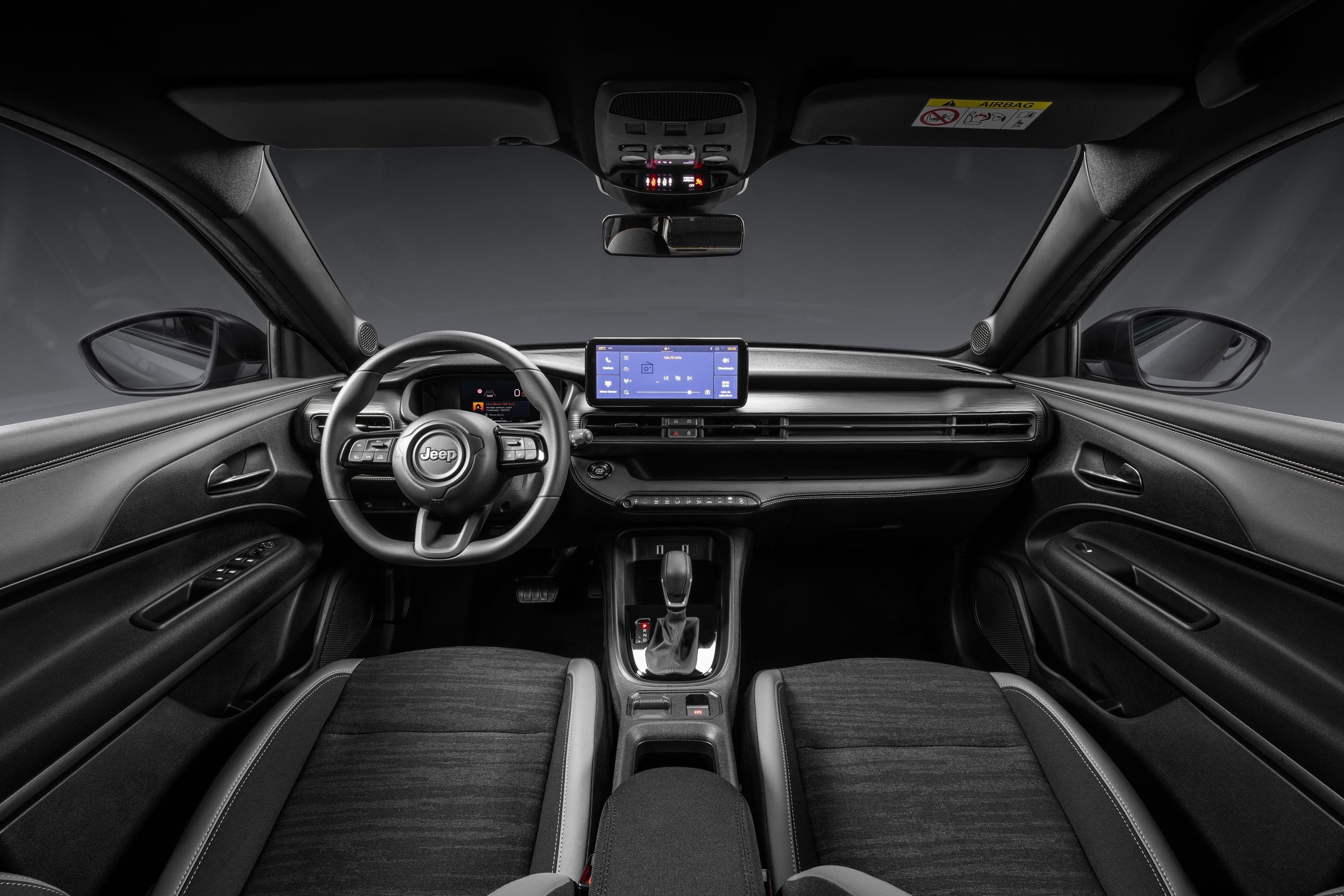 Interior de um carro Jeep moderno, com painel e estofamento em tons escuros. Volante multifuncional com o logo Jeep, tela multimídia central, saídas de ar horizontais e console com alavanca de câmbio. Os bancos dianteiros são de tecido cinza escuro com detalhes em couro.