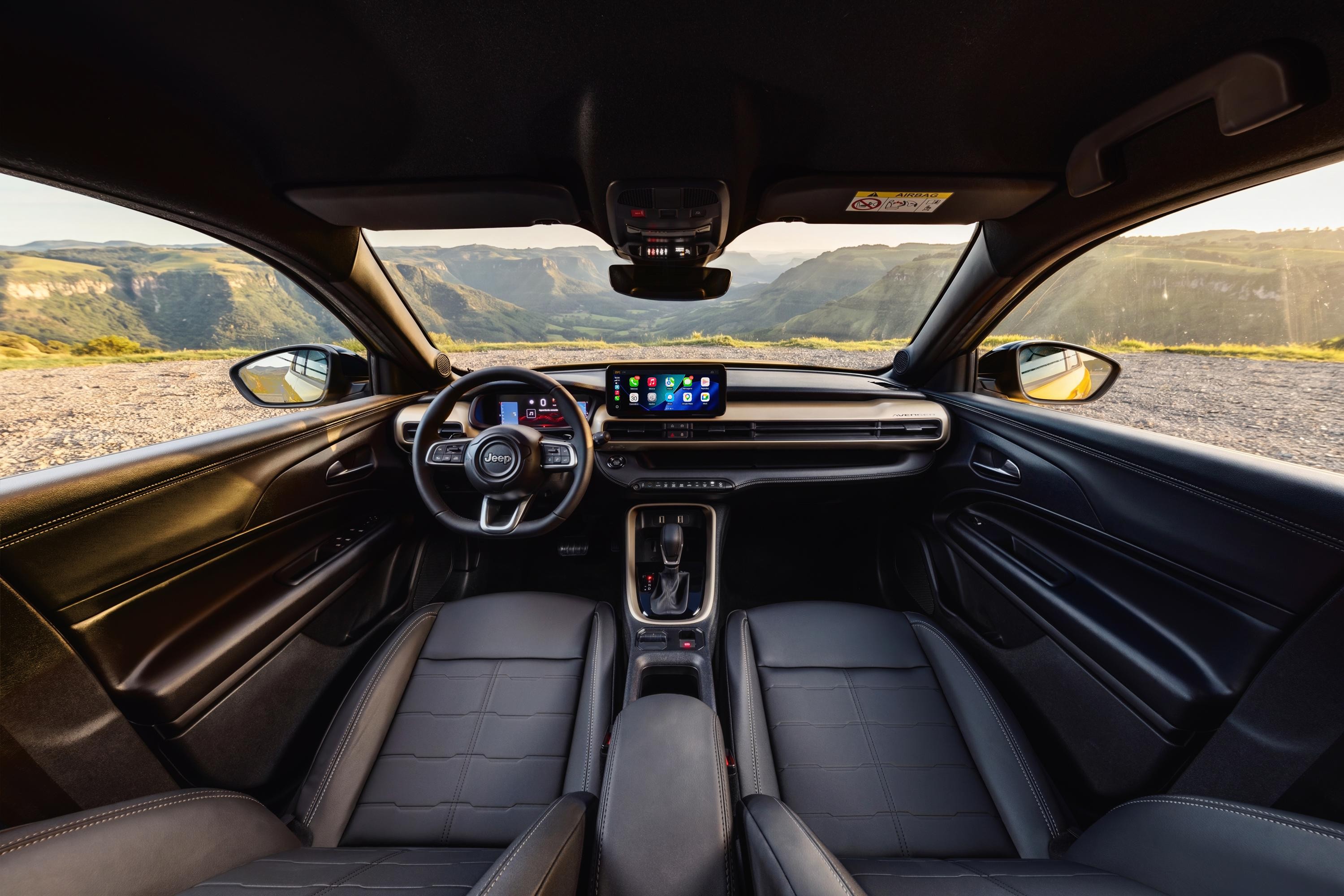 Interior de um carro Jeep preto, com bancos de couro, volante multifuncional e tela multimídia central exibindo ícones de aplicativos. O painel tem detalhes em tom dourado. A vista através do para-brisa e das janelas laterais mostra uma paisagem montanhosa com céu claro e sol poente, iluminando a estrada de terra. Os retrovisores externos refletem a paisagem.