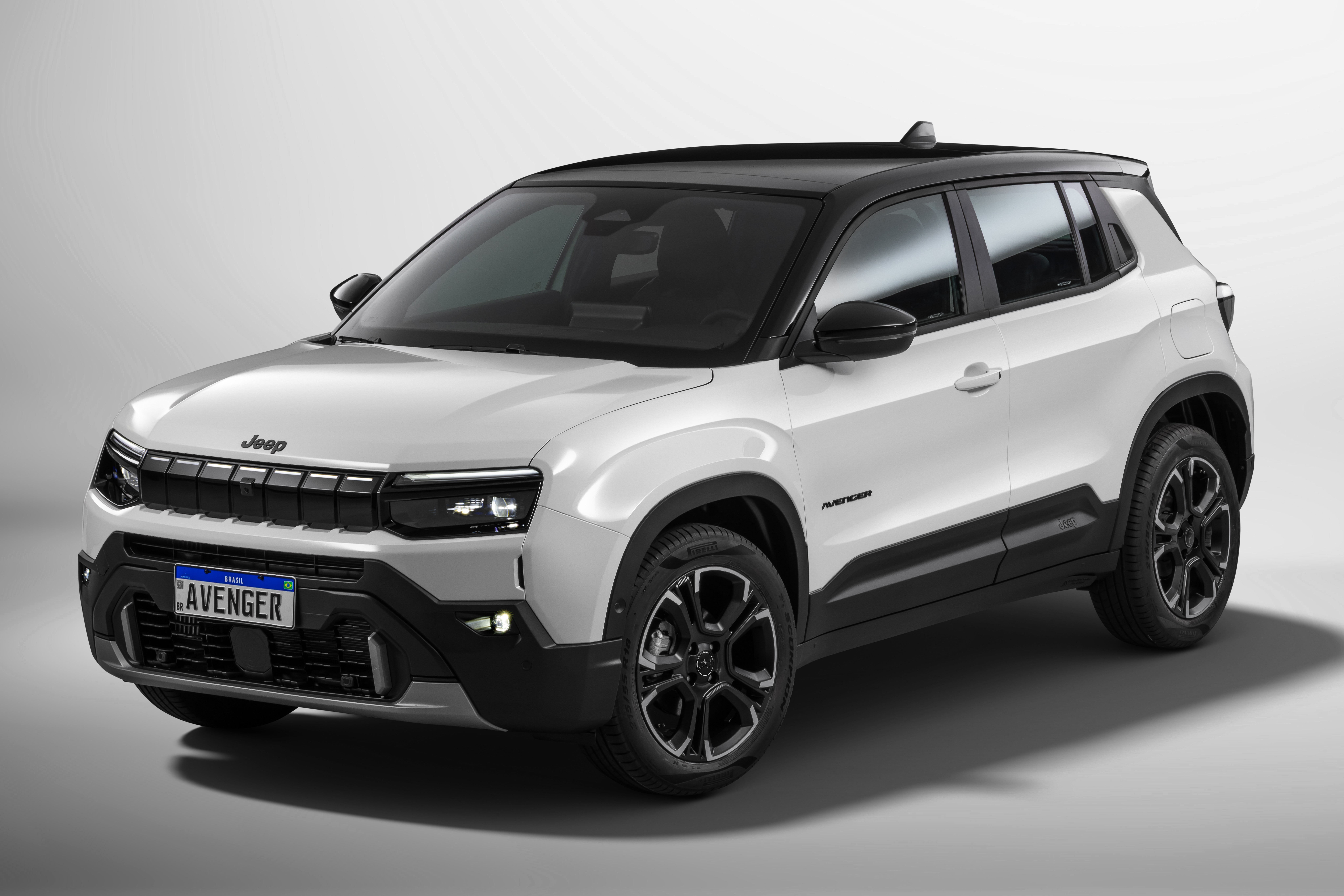 SUV Jeep Avenger branco com teto preto, visto de frente e lateral direita, em estúdio com fundo cinza claro. O carro tem grade frontal preta, faróis de LED acesos e placa AVENGER azul e branca. Rodas pretas com detalhes prateados e molduras dos para-lamas em plástico preto.