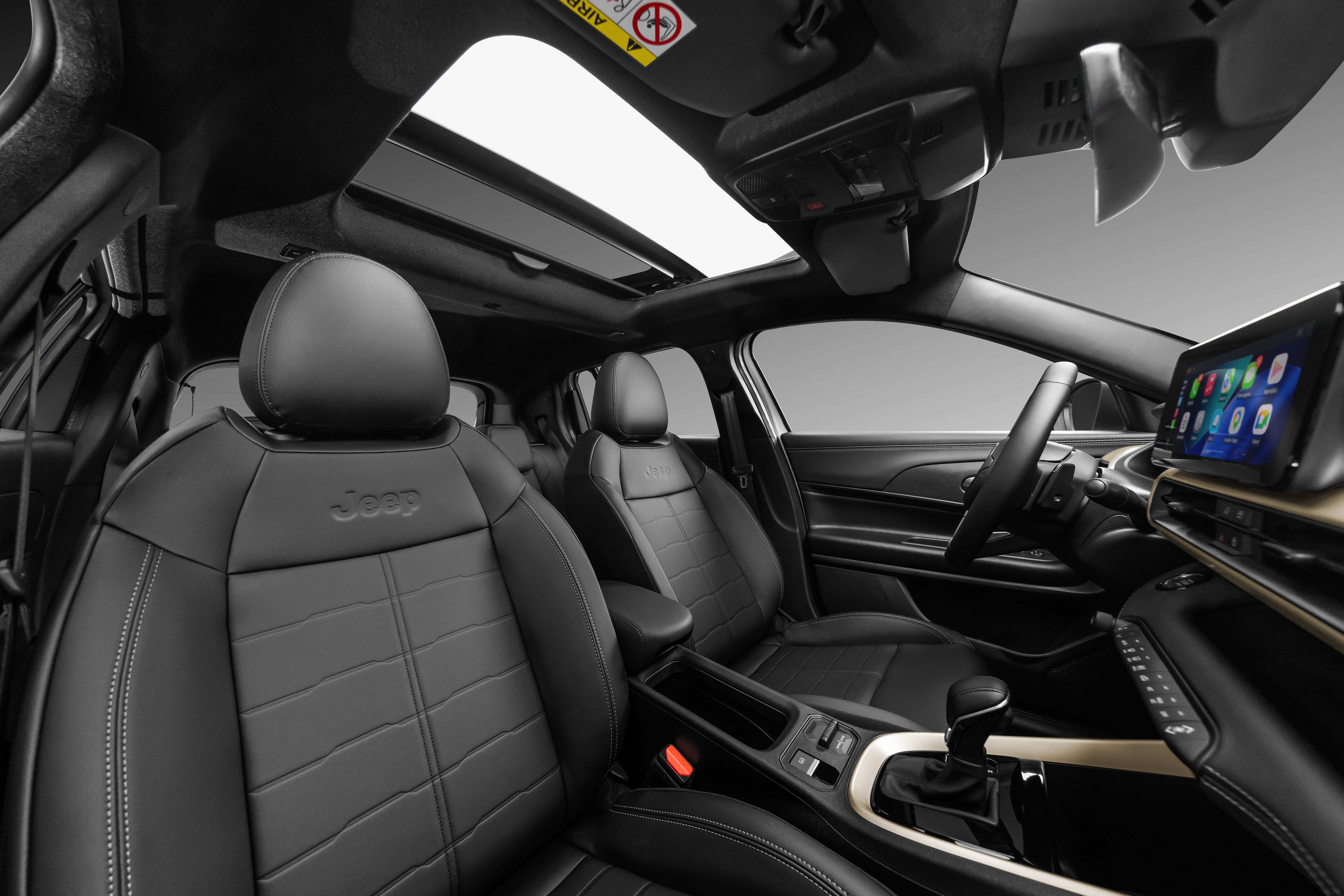 Interior de um carro moderno com bancos de couro preto, teto solar panorâmico e painel com tela multimídia. O logo Jeep está gravado nos encostos dos bancos dianteiros. O console central tem acabamento em tom claro e câmbio automático.