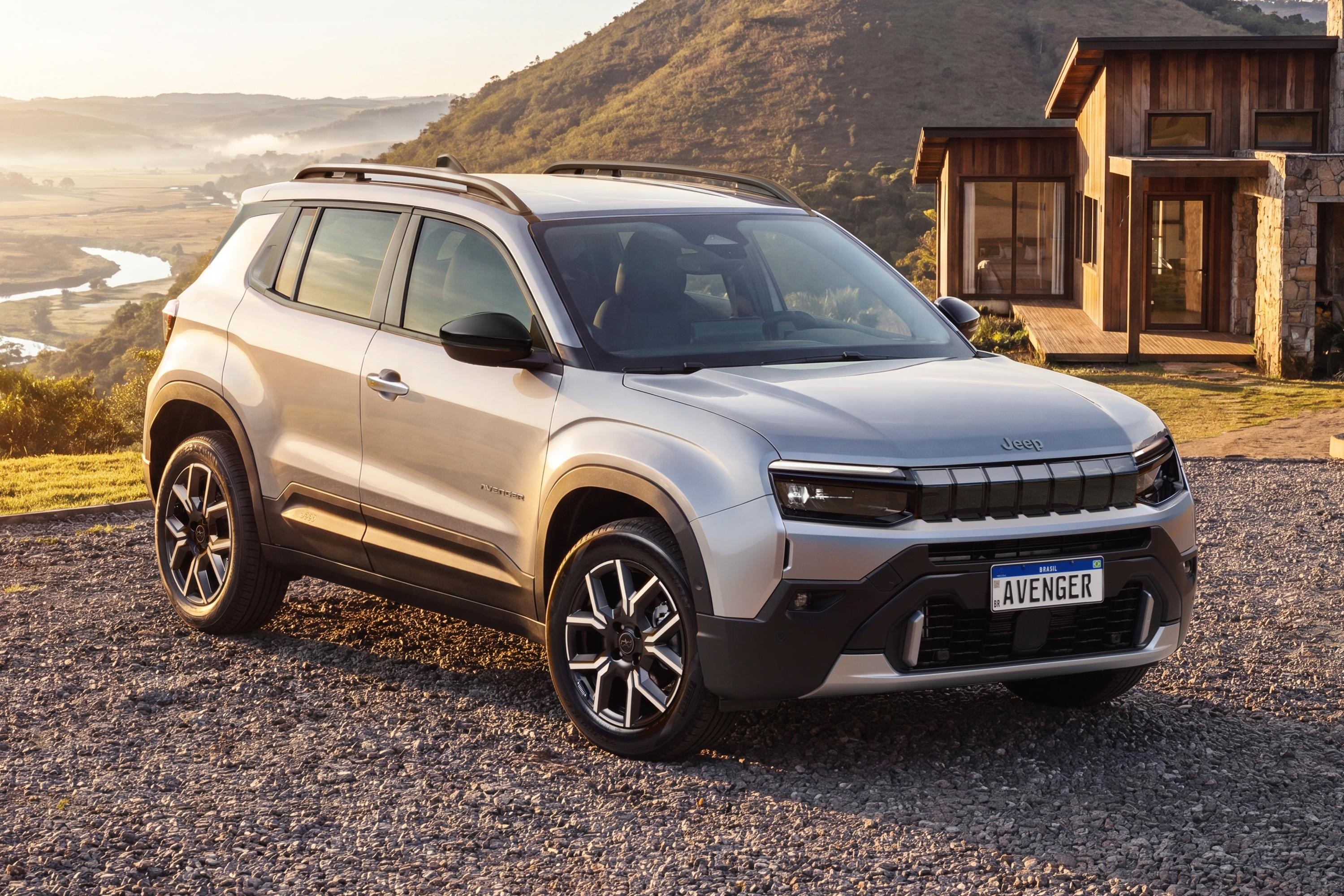 SUV Jeep Avenger prateado estacionado em estrada de cascalho, com casa de madeira e pedra ao fundo. À esquerda, um vale com rio sinuoso e montanhas sob o sol da manhã