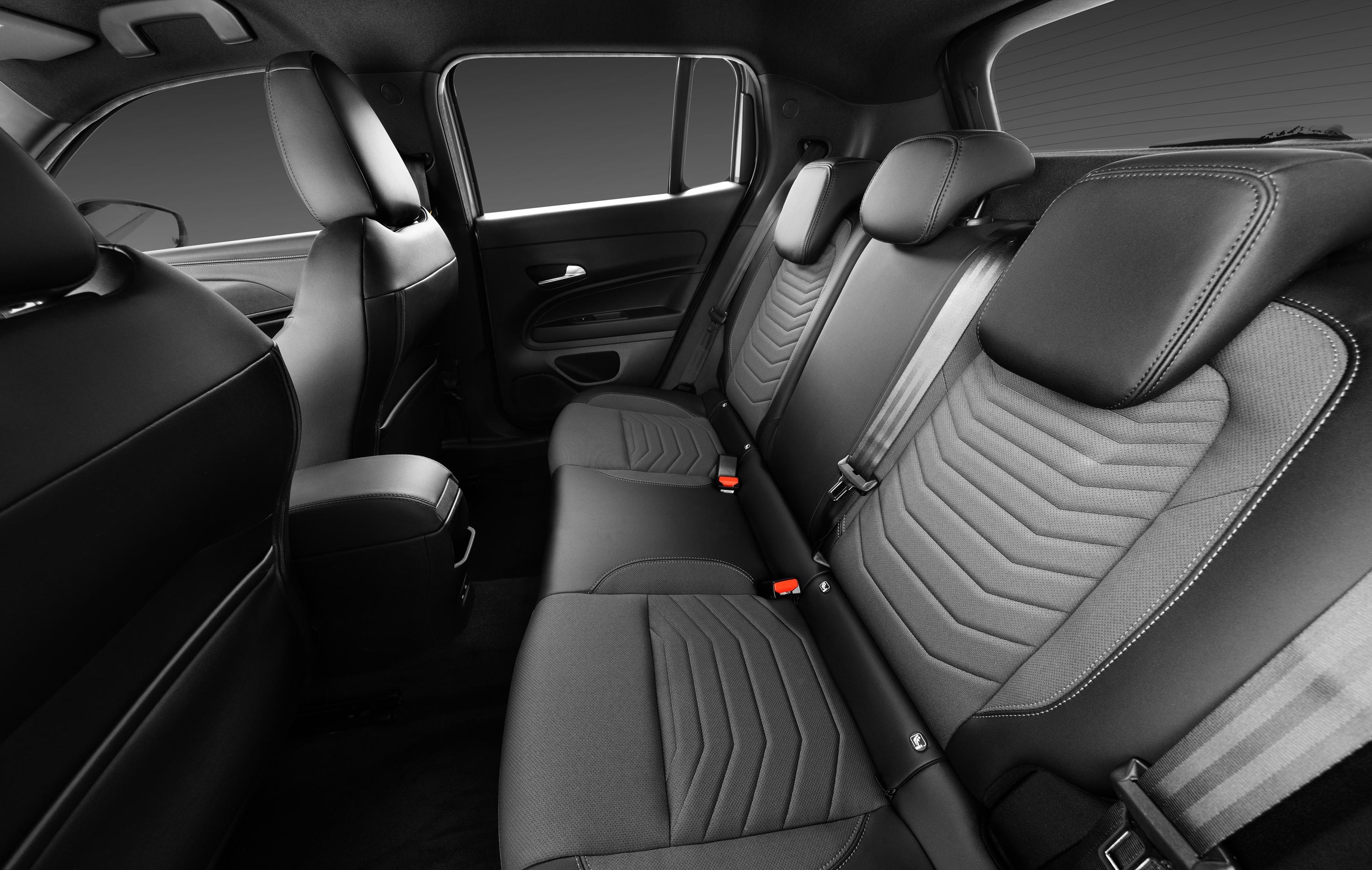 Interior de um carro com bancos traseiros em couro preto e tecido cinza escuro, com costuras brancas e padrão geométrico em zigue-zague. Os cintos de segurança estão visíveis, e o banco do passageiro dianteiro é parcialmente visível à esquerda