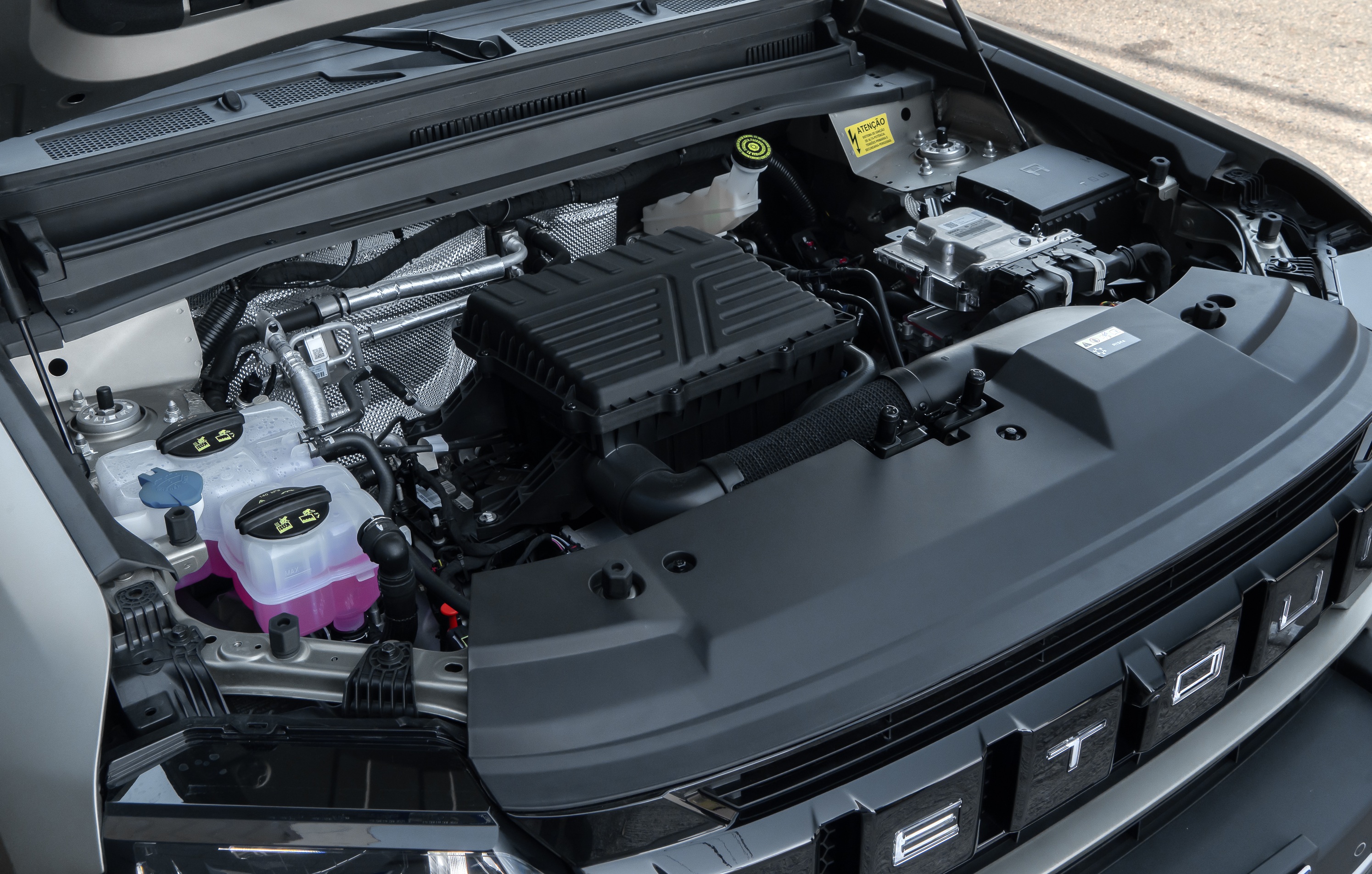 Compartimento do motor de um carro com o capô aberto, mostrando o motor, reservatórios de fluidos e componentes eletrônicos. O motor tem uma tampa preta texturizada, e há dois reservatórios transparentes com tampas pretas e um com tampa azul, um deles com líquido roxo. O fundo é cinza claro.
