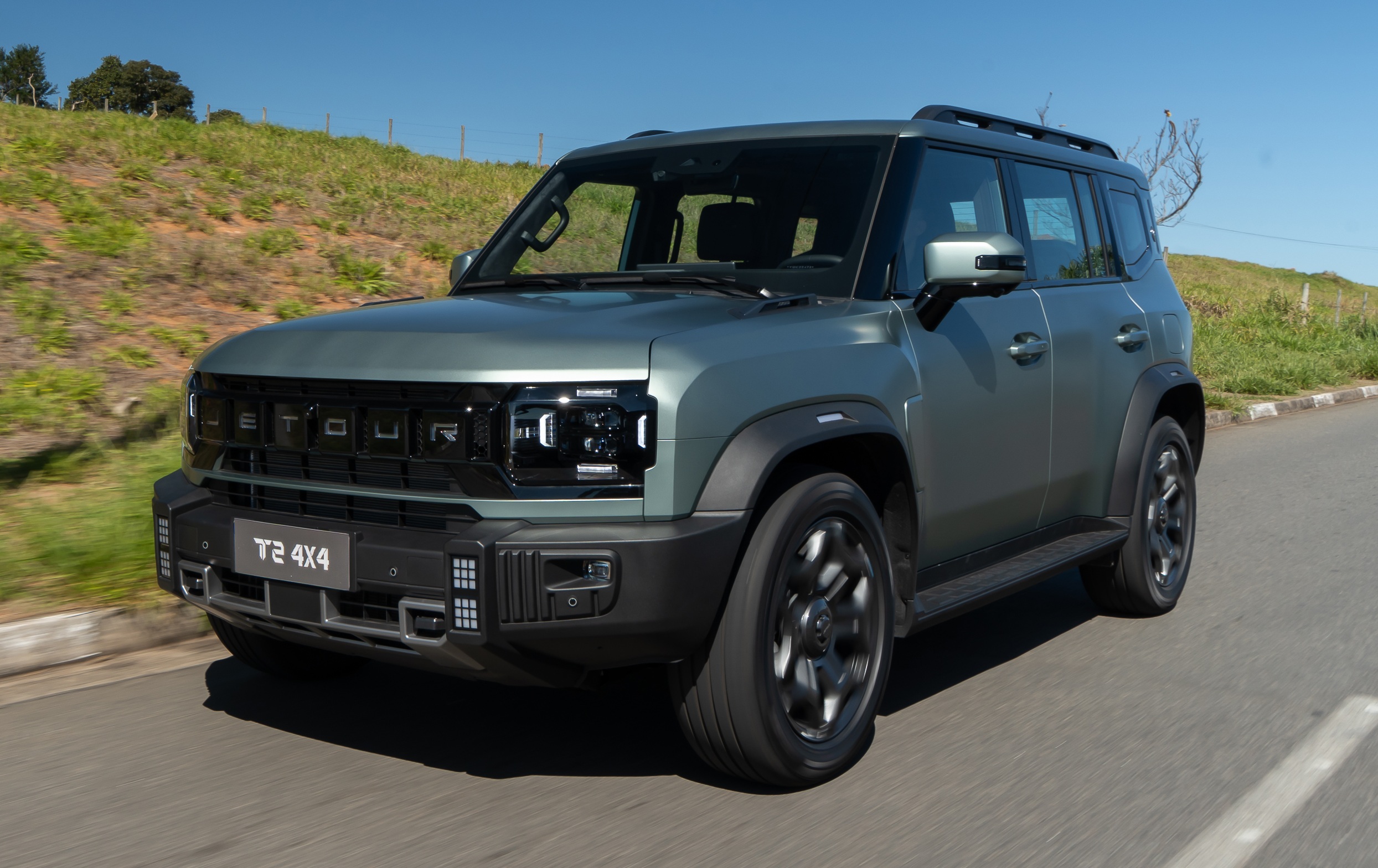 SUV Jetour T2 4x4 cinza-esverdeado em movimento, visto de frente e lateral, em uma estrada asfaltada com gramado e céu azul ao fundo