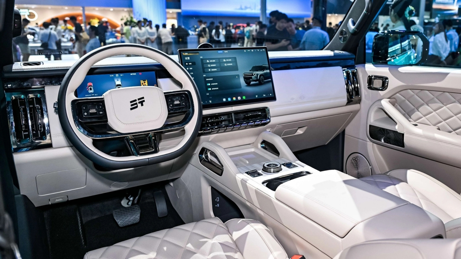 Interior de um carro moderno com acabamento em tons claros, volante branco e preto, painel digital e tela multimídia exibindo um SUV. Bancos e portas com estofamento acolchoado em losangos, e detalhes cromados no console central. Pessoas desfocadas ao fundo sugerem um evento ou feira