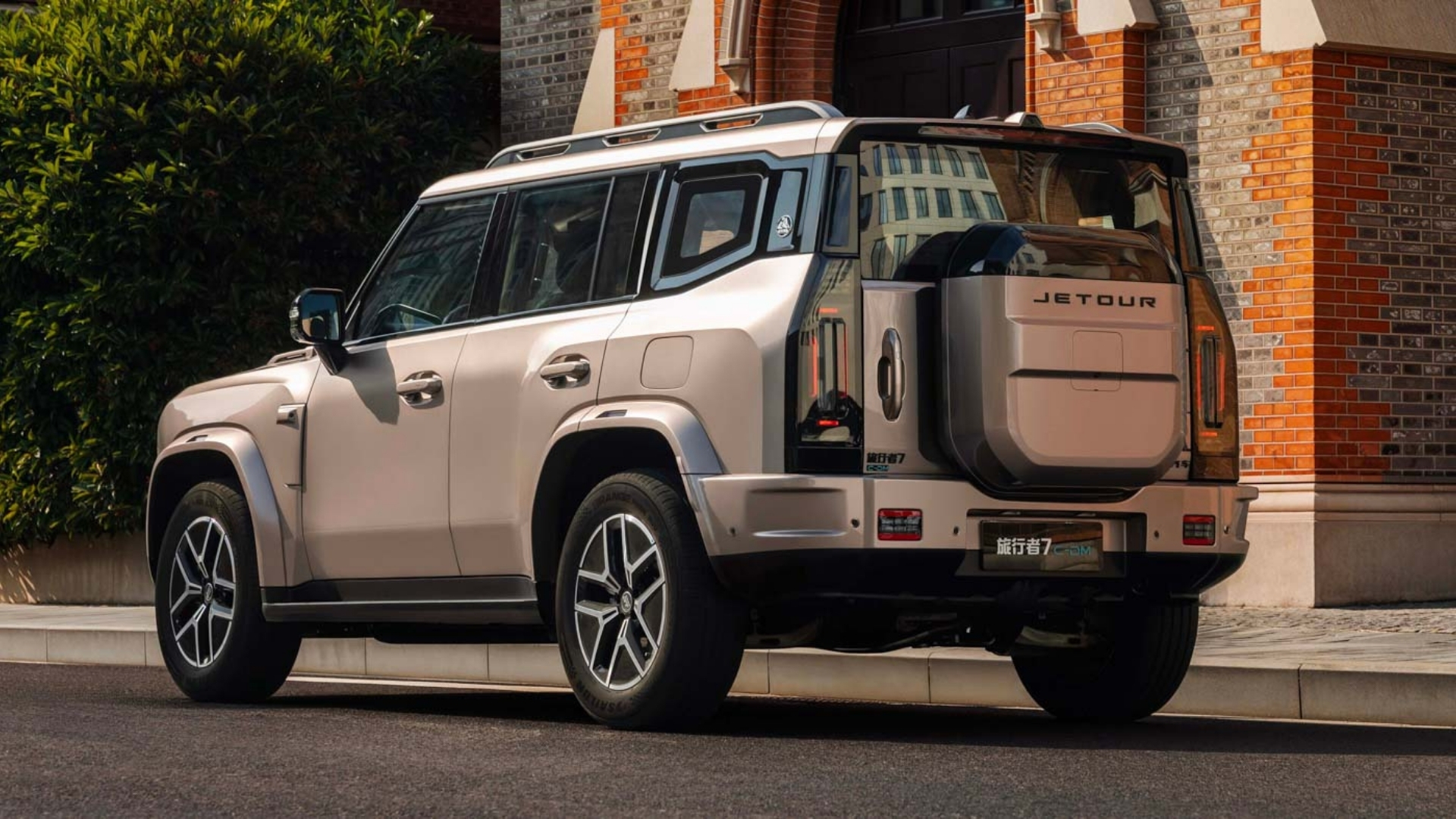 SUV Jetour Traveller bege claro estacionado em uma rua, com a traseira e lateral direita visíveis. Ao fundo, um prédio de tijolos e folhagem verde