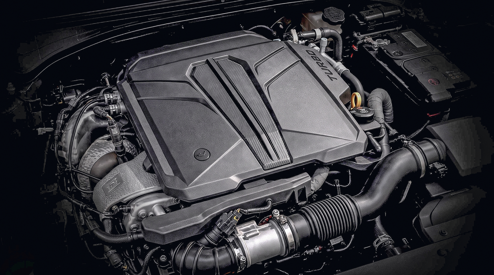 Motor de carro moderno com tampa cinza escura e detalhes em fibra de carbono, exibindo a palavra TURBO em relevo. Componentes como mangueiras, fios e a bateria são visíveis ao redor, em um ambiente escuro que destaca o motor