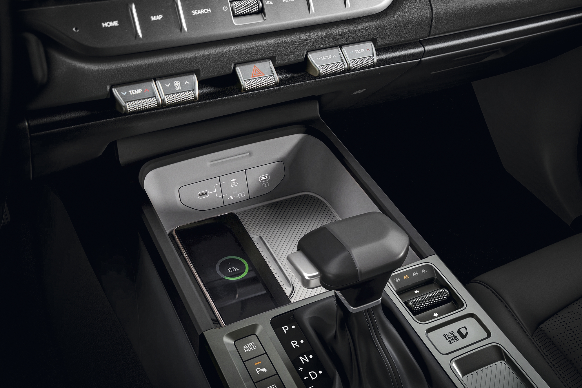 Interior de um carro com console central cinza e preto. Um smartphone preto carrega sem fio em um compartimento, exibindo 88% de bateria. Acima, botões de controle de temperatura e alerta. À direita, a alavanca de câmbio automática e outros controles do veículo