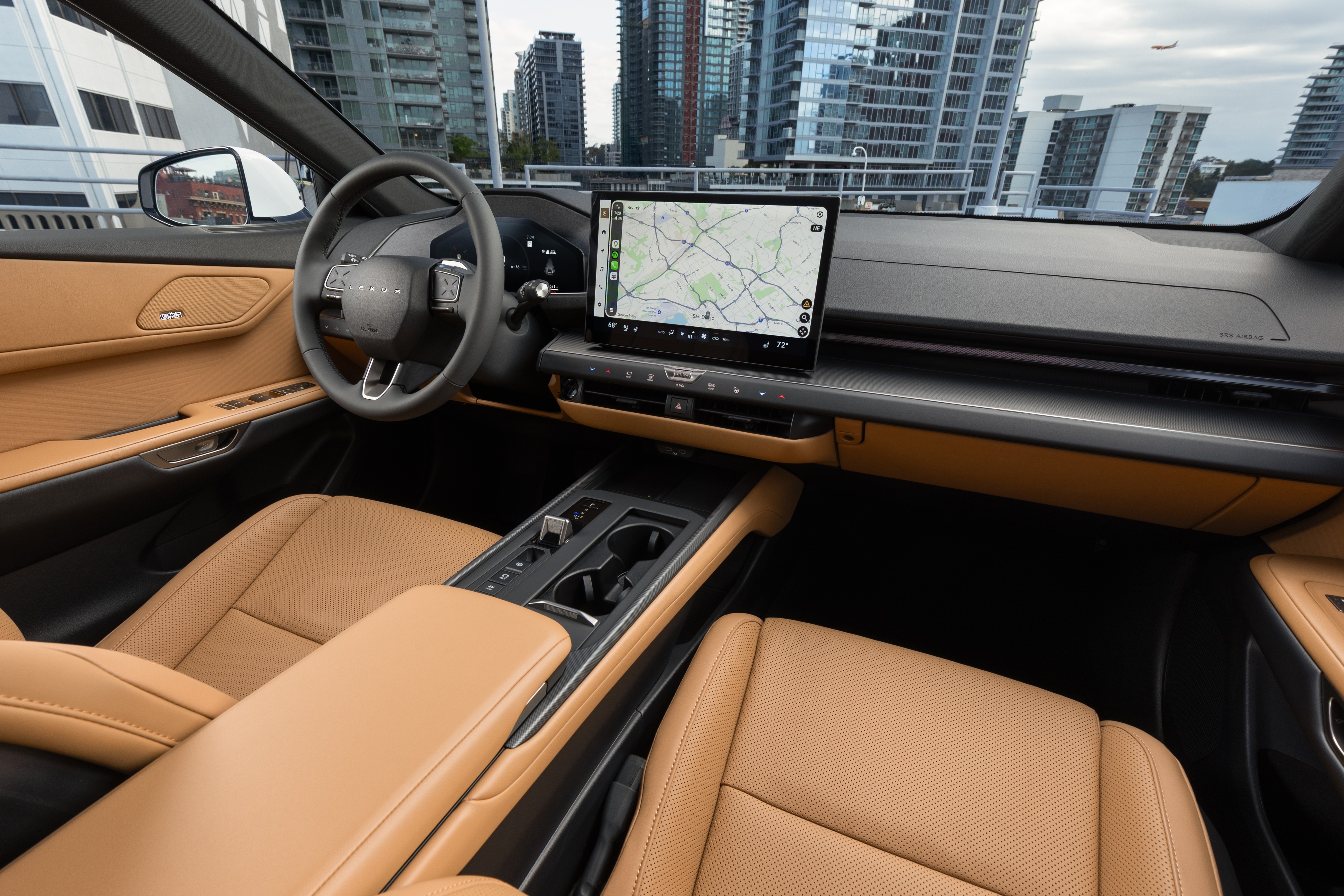 Interior de um carro moderno com bancos de couro marrom-claro e detalhes pretos. O painel possui uma tela grande exibindo um mapa de navegação. Ao fundo, prédios altos e um céu nublado com um avião distante