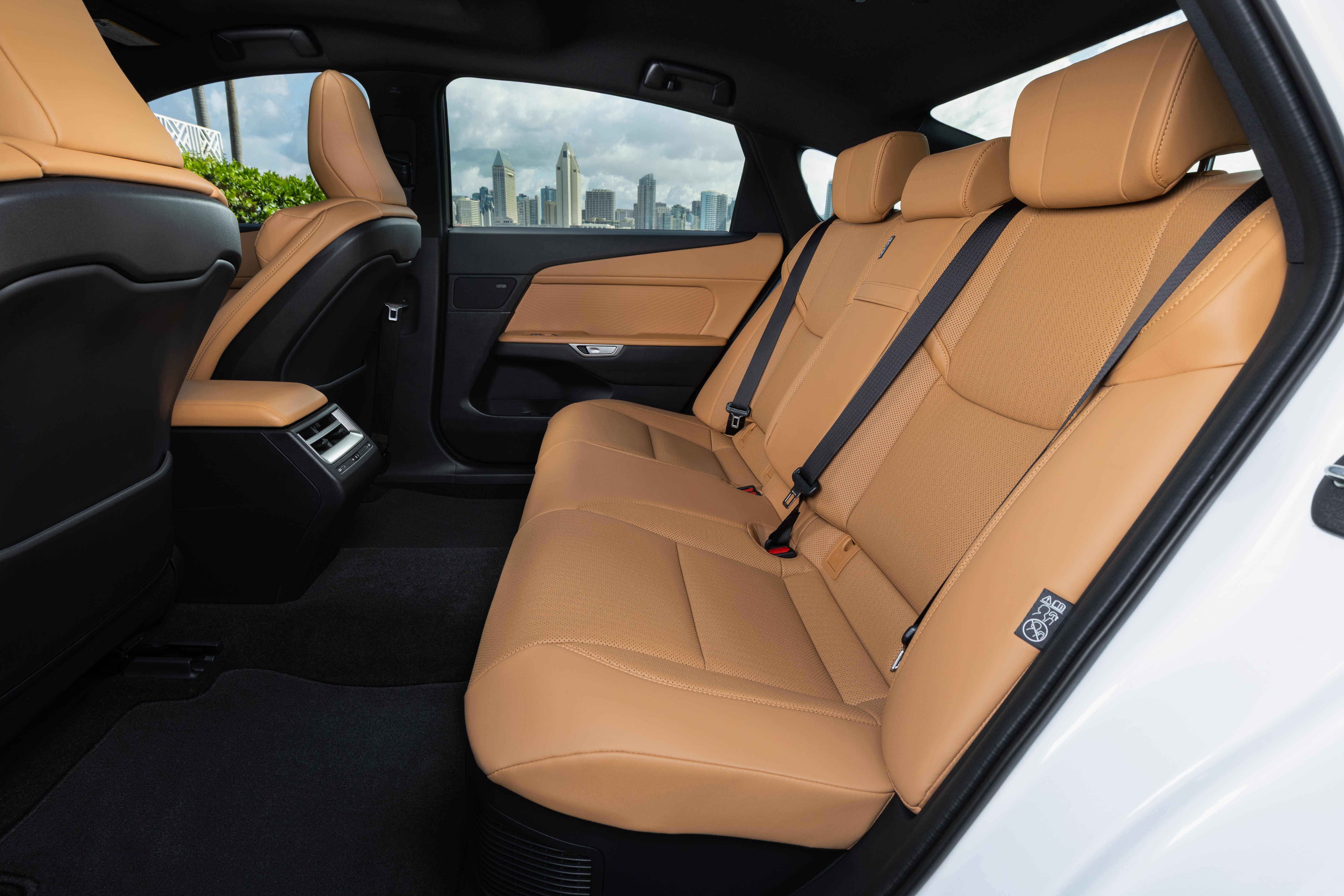 Interior de carro com bancos traseiros de couro marrom-claro e cintos de segurança pretos. O piso é forrado com carpete preto. Pela janela, vê-se um horizonte urbano com prédios altos sob céu nublado