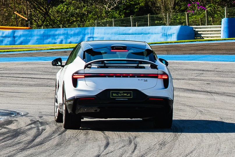 Traseira de um carro branco Lotus Eletre em uma pista de corrida, com faixas azuis e amarelas, e vegetação ao fundo