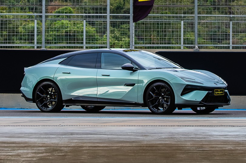 Carro esportivo elétrico Lotus Emeya azul-claro metálico com teto preto, rodas pretas e detalhes pretos, parado em uma pista de corrida com grade e árvores ao fundo