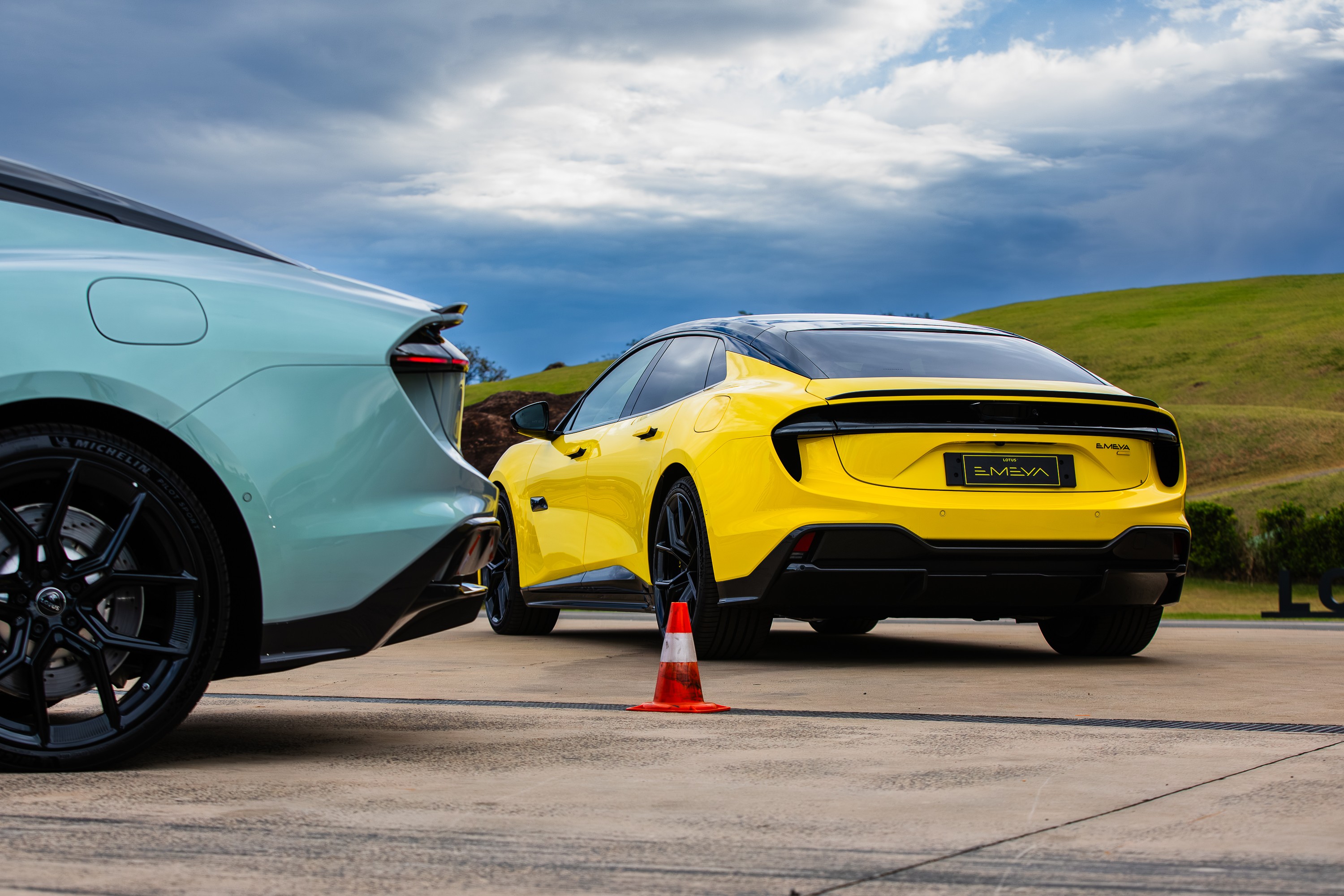 Dois carros esportivos modernos, um verde-água à esquerda e um amarelo à direita, estacionados em uma pista de asfalto. O carro amarelo tem a placa EMEVA e um cone de trânsito laranja à frente. Ao fundo, colinas verdes e um céu nublado com nuvens escuras