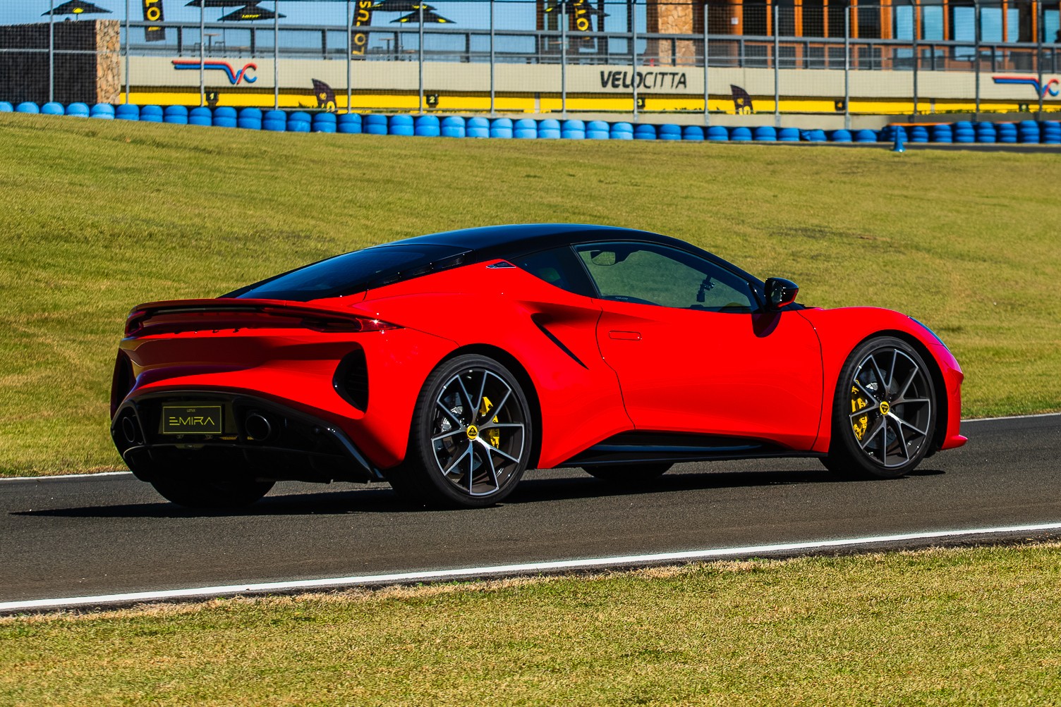 Carro esportivo vermelho com teto preto, modelo Lotus Emira, parado em uma pista de corrida. Ao fundo, uma barreira de pneus azuis e uma construção com a palavra VELOCITTA