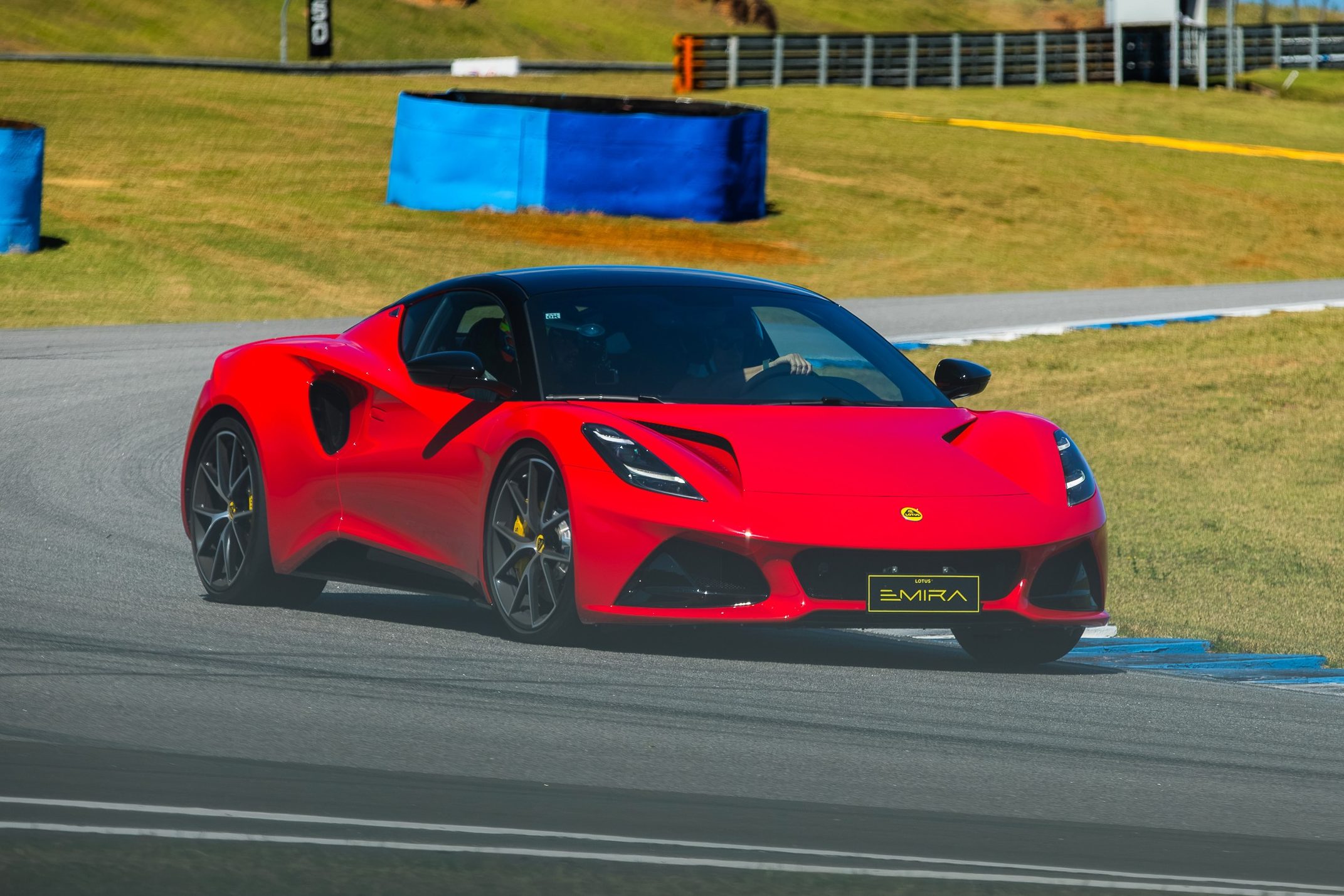 Carro esportivo Lotus Emira vermelho com teto preto, placa EMIRA e rodas escuras com pinças amarelas, em uma pista de corrida cinza com faixas brancas e azuis, gramado verde ao fundo