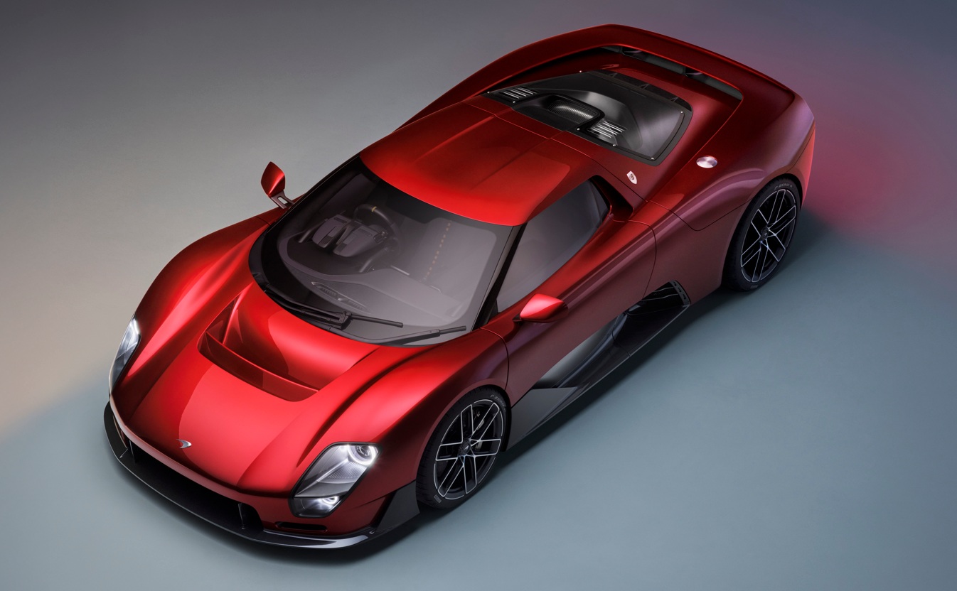 Carro esportivo vermelho metálico, visto de cima, com design aerodinâmico e detalhes em preto fosco, como as rodas e a parte inferior da carroceria. O veículo está parado sobre um fundo cinza-azulado, com iluminação suave destacando suas curvas.