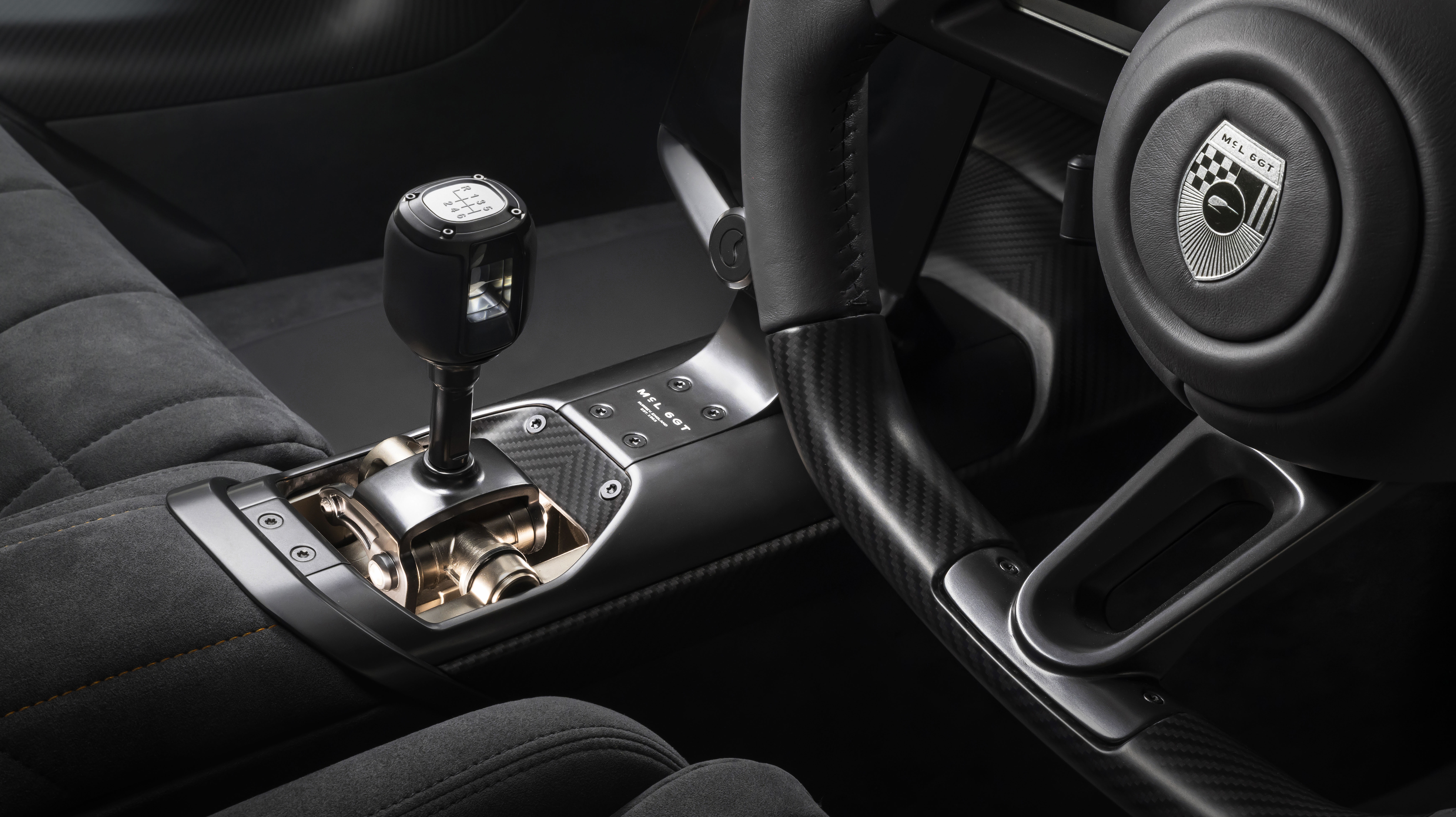 Interior de carro esportivo com câmbio manual de seis marchas e ré, alavanca preta e base dourada exposta. Volante multifuncional com acabamento em fibra de carbono e emblema Mil.ot no centro. Bancos em alcântara cinza com costura laranja. Detalhes em preto e prata, criando um ambiente sofisticado e tecnológico